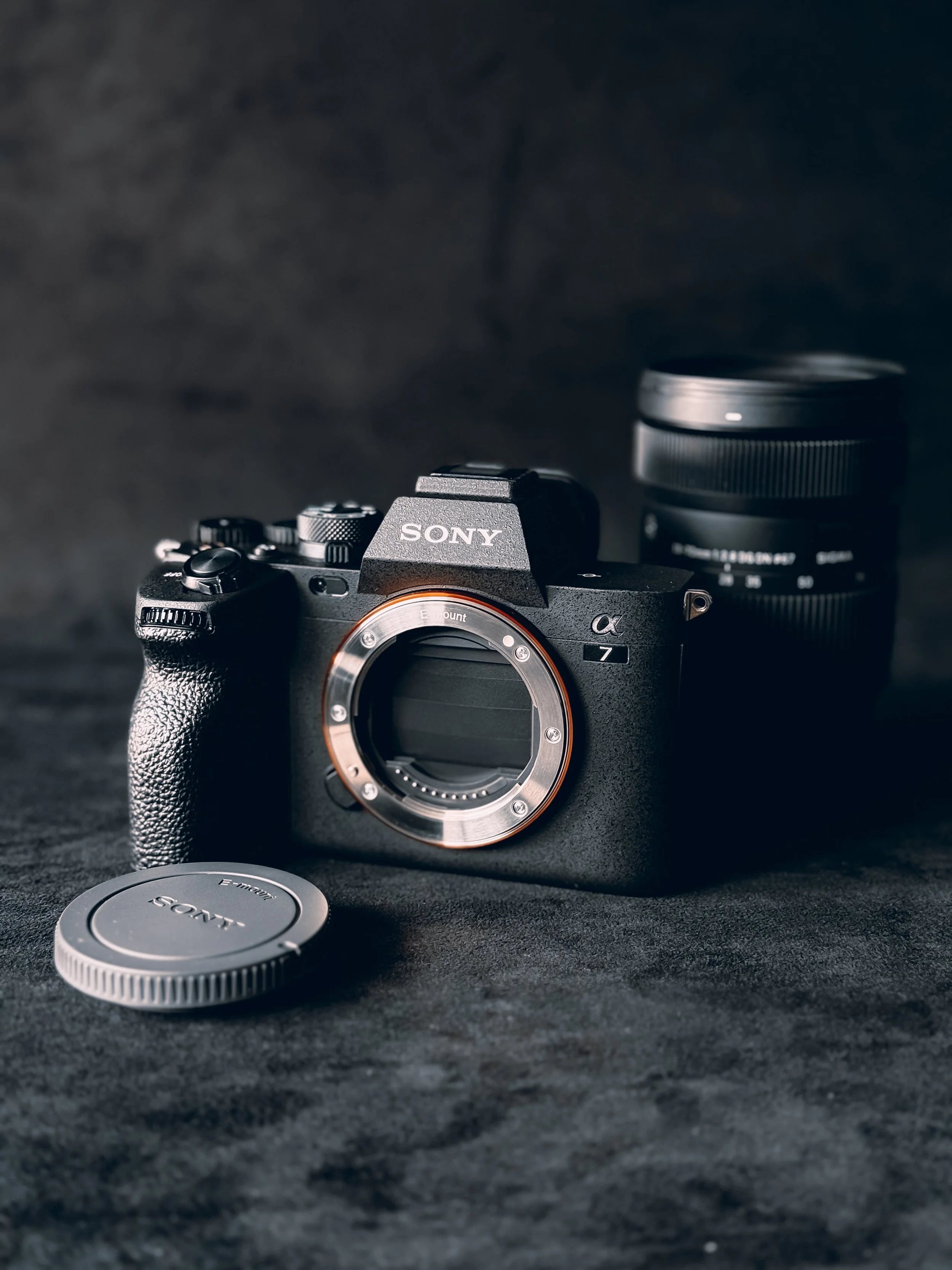 A Sony mirrorless camera body without a lens attached, placed on a dark surface, with a camera lens and a lens cap nearby.