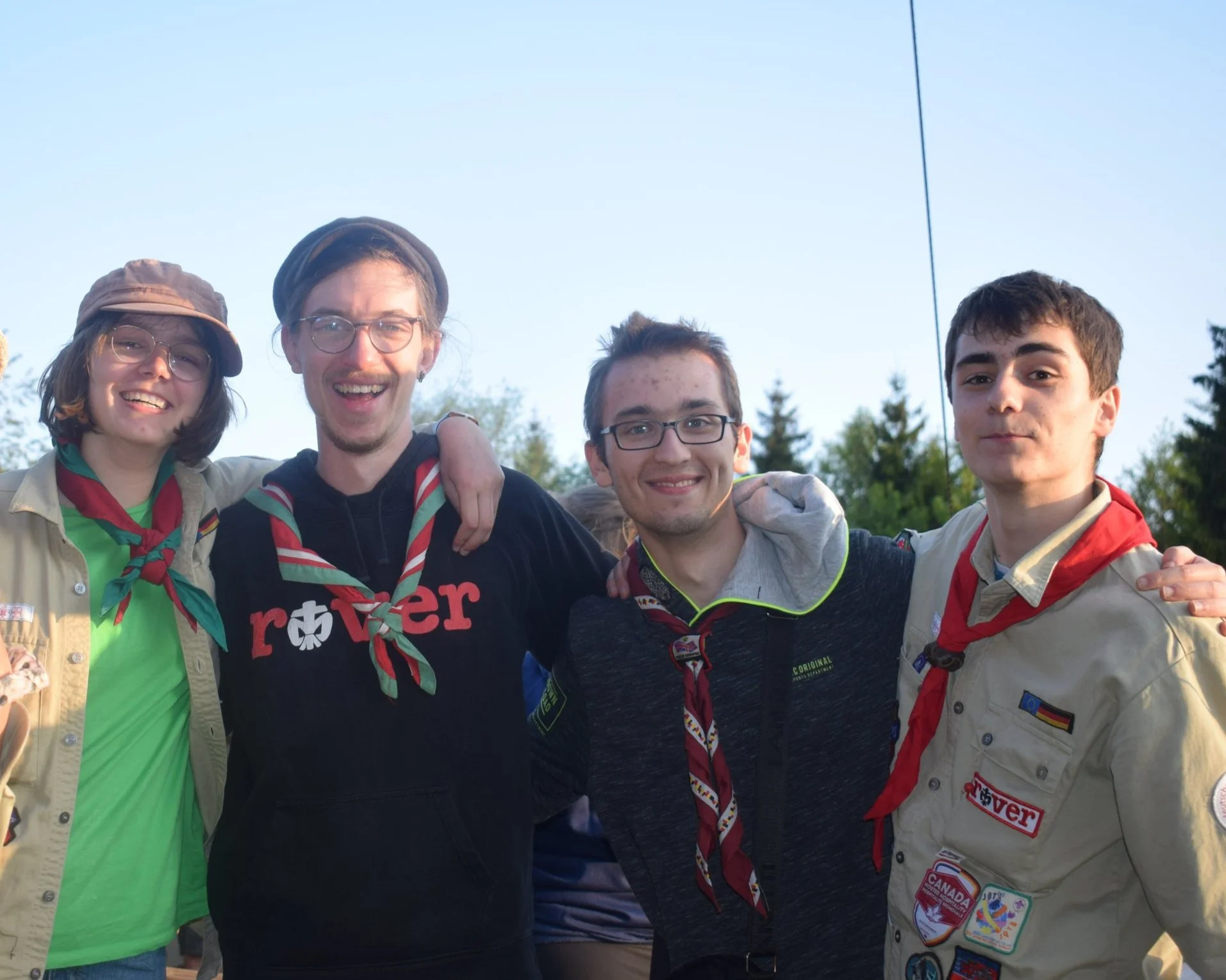 Fünf junge Menschen, die zusammen in der Natur stehen, lachen und freundlich in die Kamera schauen, bei einem Outdoor-Event oder einer Zeltlageraktivität.