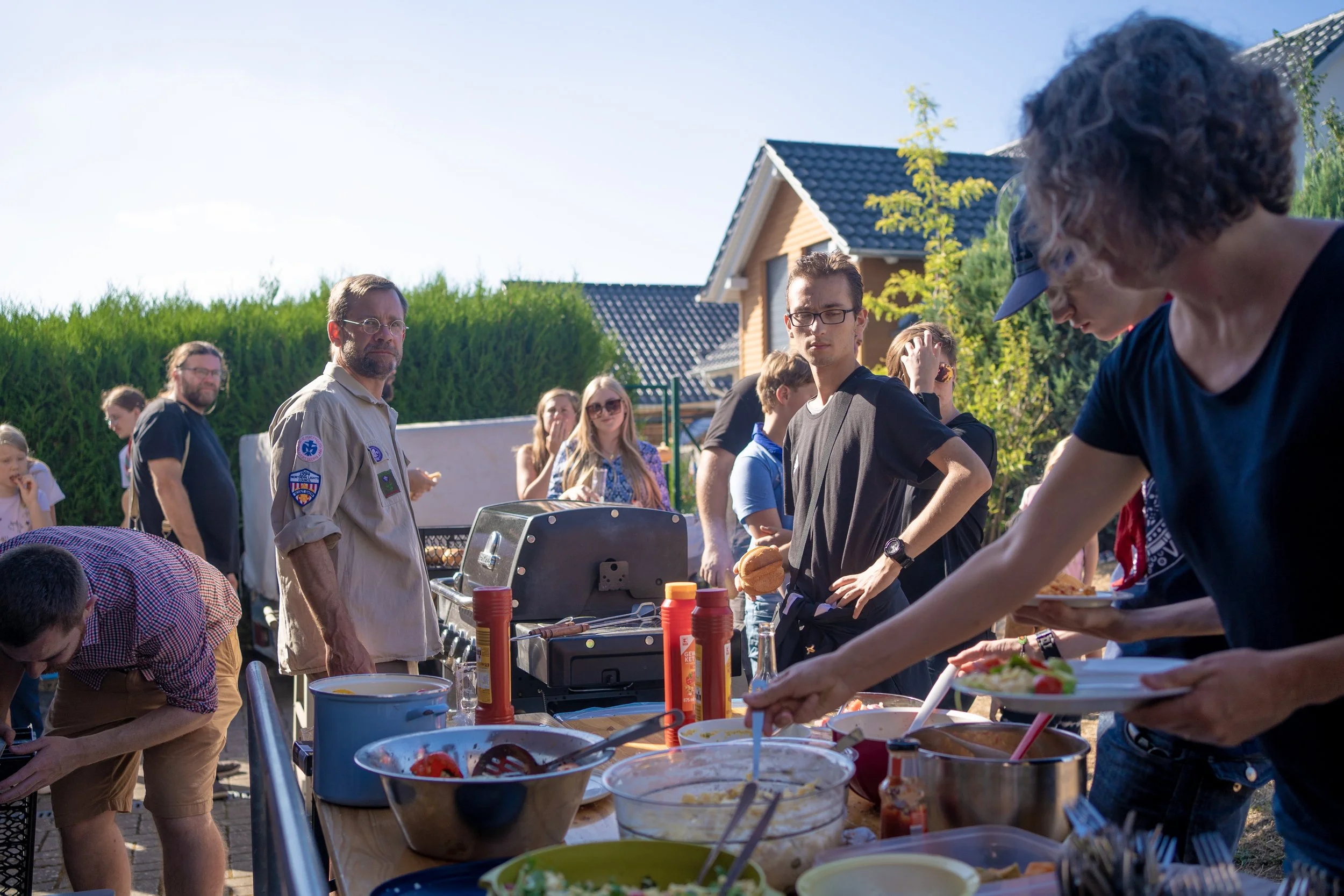 Menschen beim Grillen und Buffet im Garten an sonnigem Tag, mit Haus und Bäumen im Hintergrund.