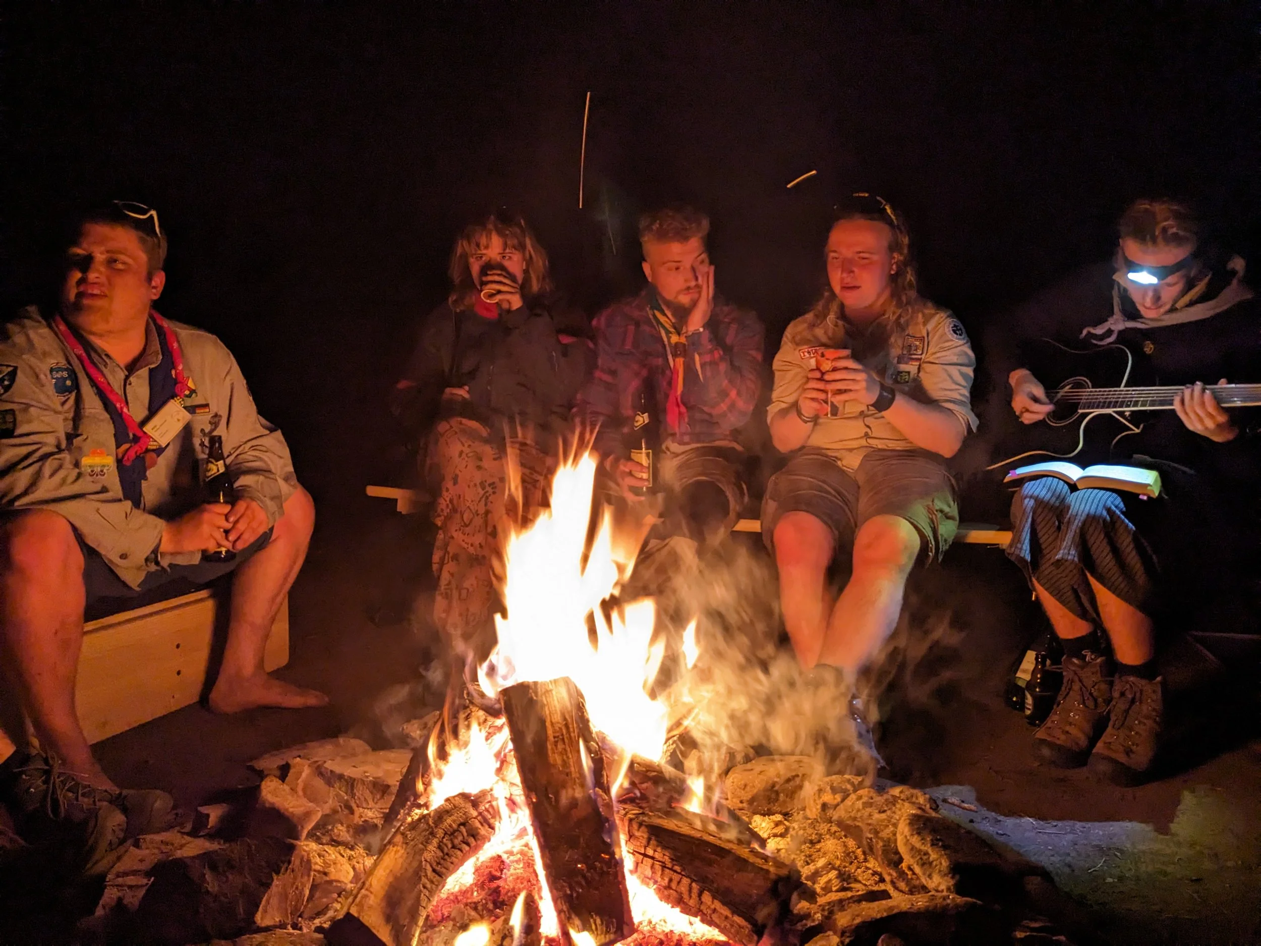Gruppe von fünf jungen Leuten sitzen um ein Lagerfeuer in der Dunkelheit, einige spielen Gitarre, andere singen und entspannen sich.