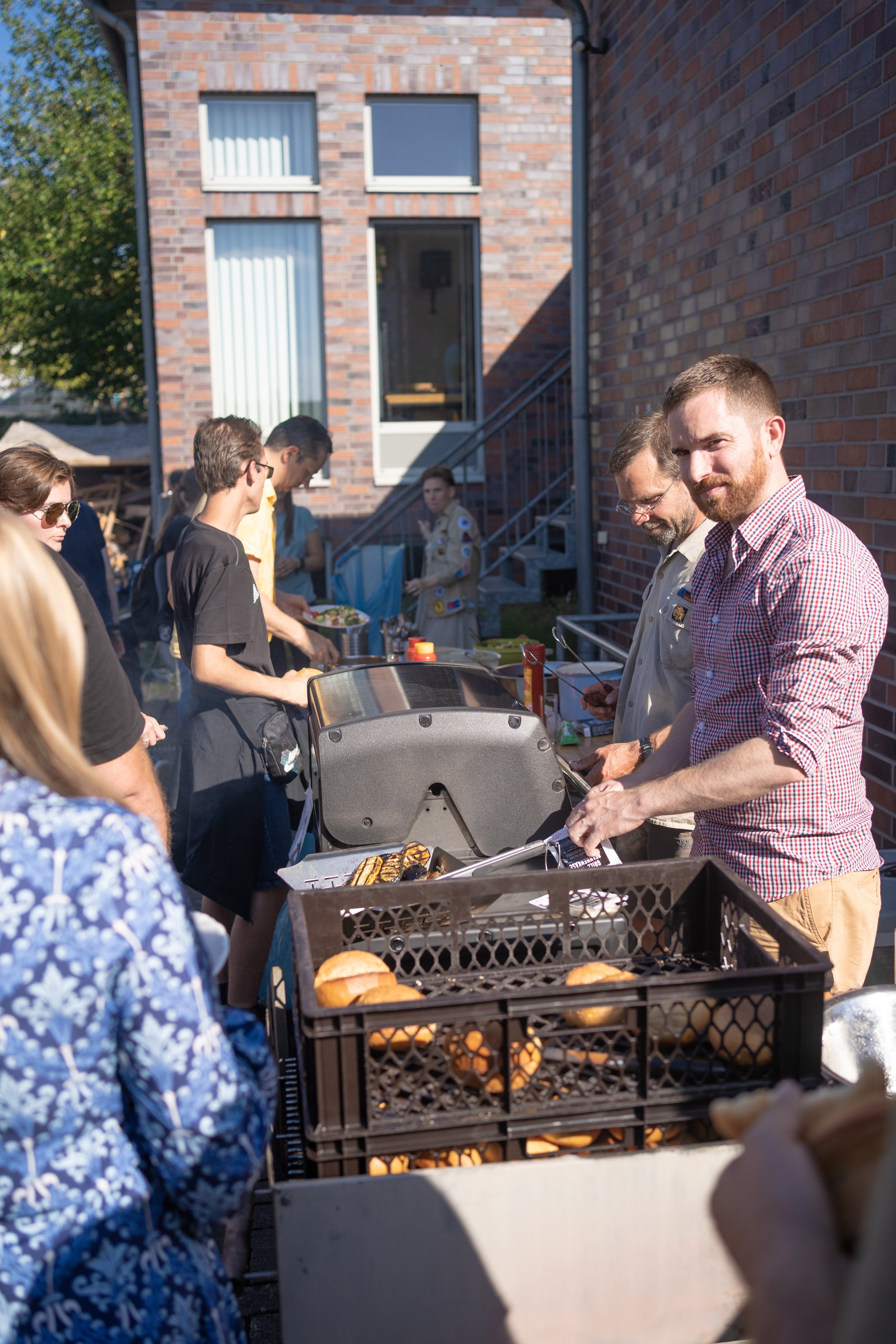 Menschen beim Grillen auf einer Gartenparty, einige bestellen Hamburger, andere warten in der Schlange, im Hintergrund eine Person in Boy Scout Uniform.