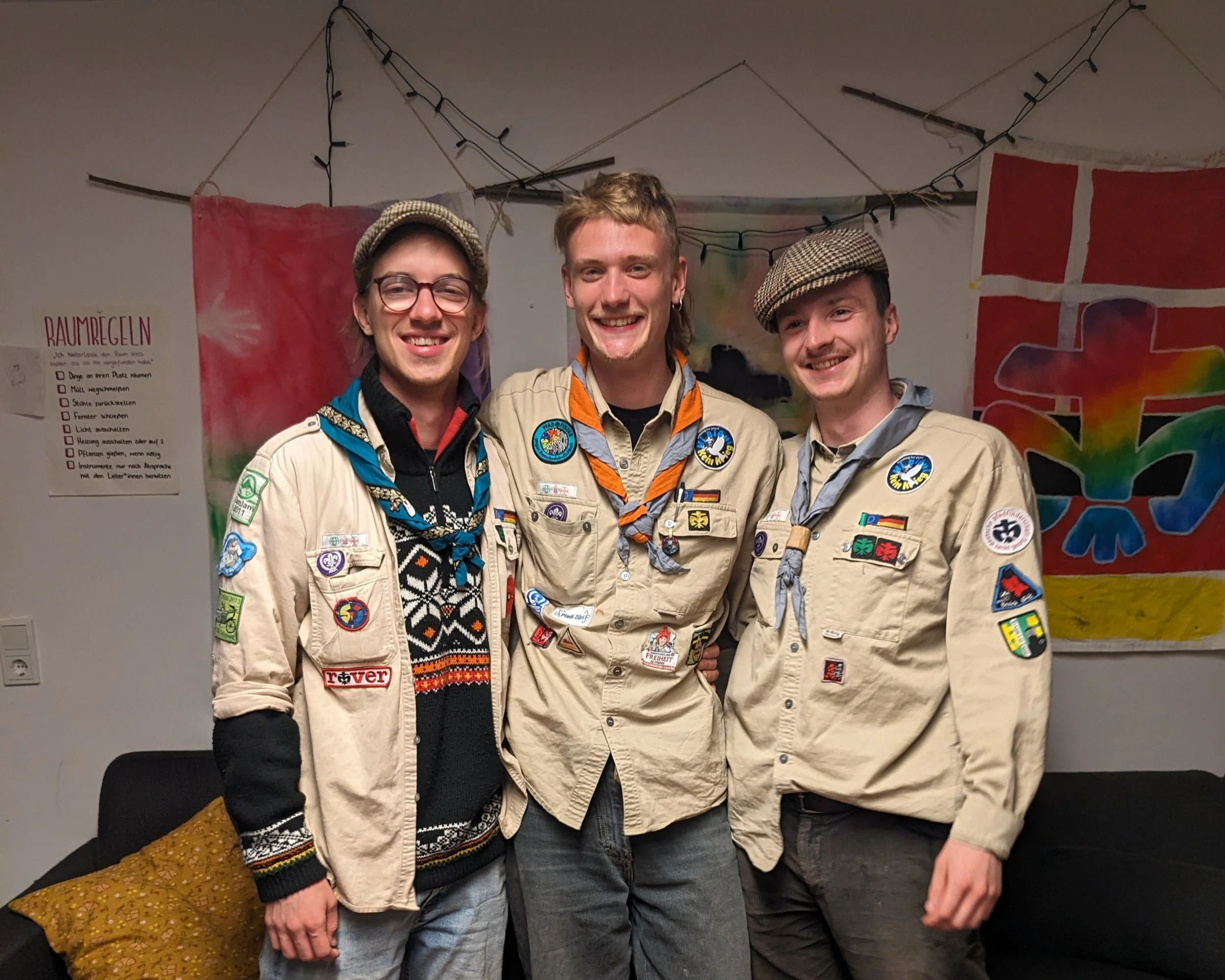 Drei junge Männer in Boy Scout-Uniformen mit Abzeichen und Halstüchern, lachend und zusammen stehend in einem Zimmer mit bunten Wanddekorationen.