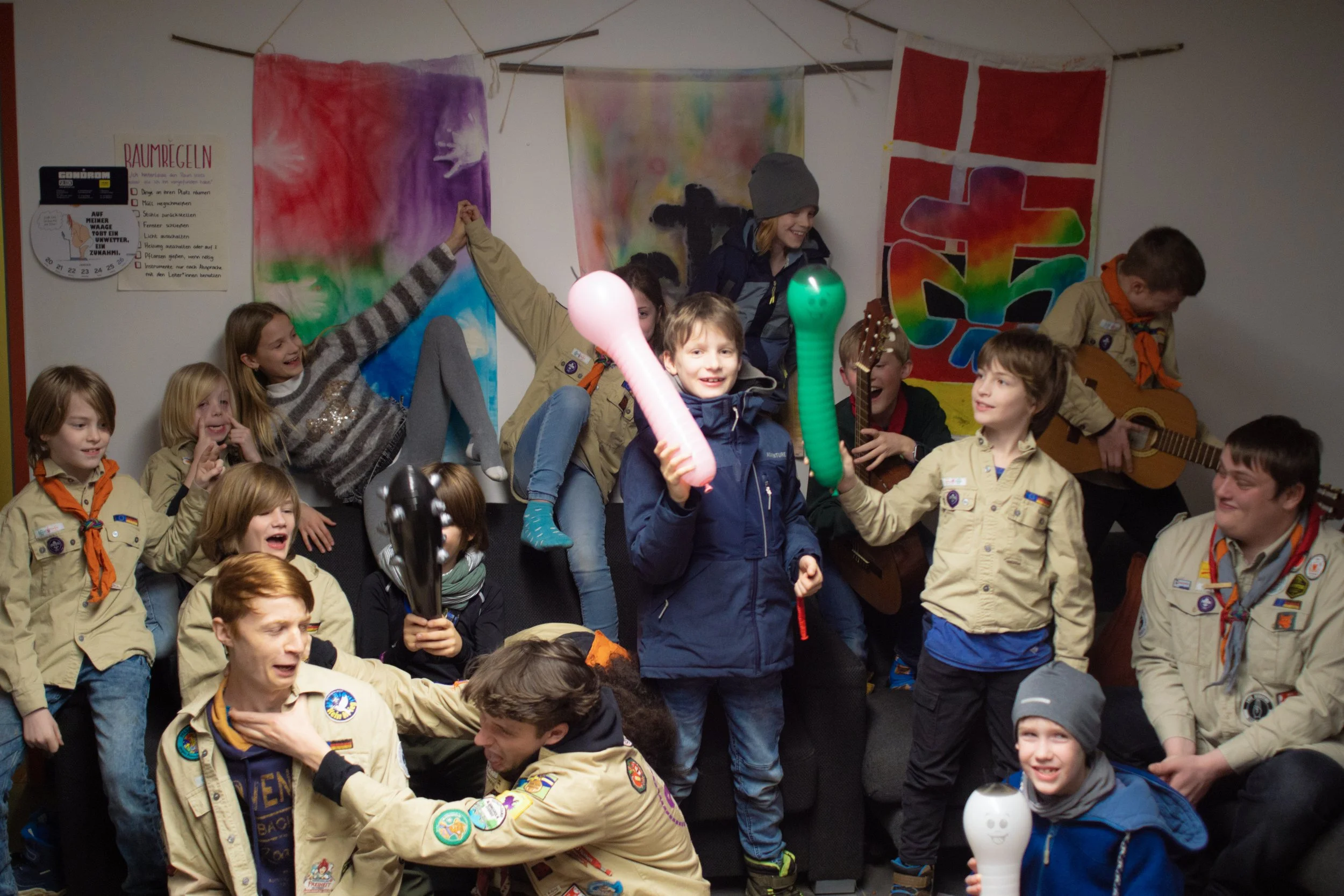 Gruppen von Jungen und Mädchen in Boy Scouts Uniformen, die in einem Raum Spaß haben, spielen und Gitarre spielen. Manche halten Luftschlangen und sonstige Luftballons.