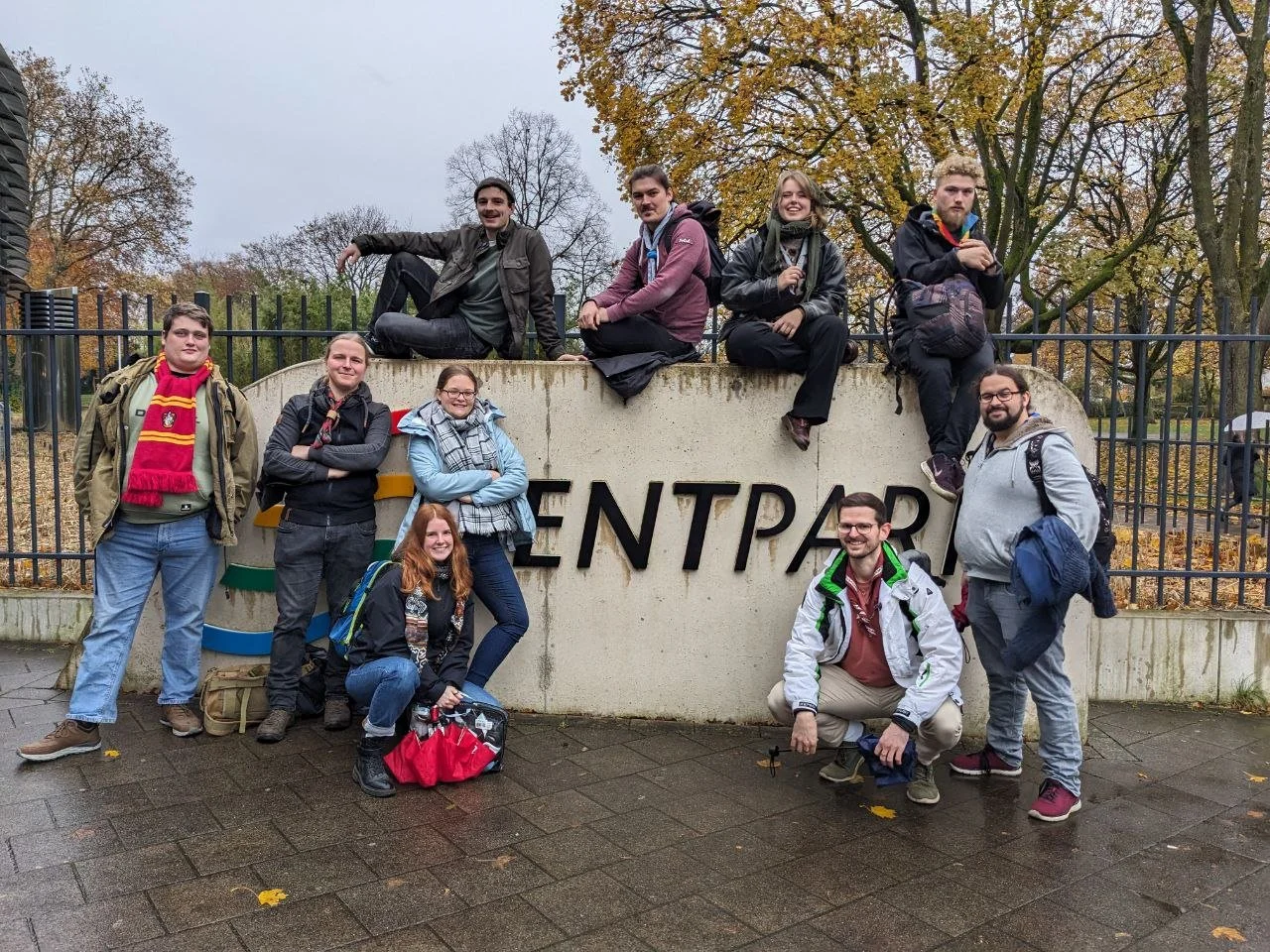 Eine Gruppe von zehn jungen Erwachsenen posiert vor dem Eingangsschild eines Parks, auf dem steht »ENTRANCE«. Sie sind lächelnd und tragen starre, wetterfeste Jacken und Rucksäcke, während sie in einem herbstlichen Park sind, mit bunten Blättern und Bäumen im Hintergrund.