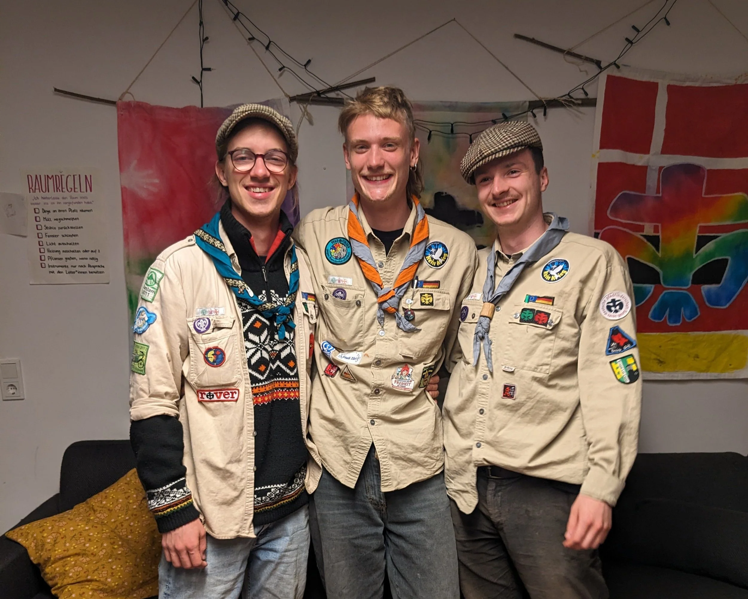Drei junge Männer in Scouts-Uniformen stehen nebeneinander und lächeln in einem Zimmer mit bunten Wanddekorationen.
