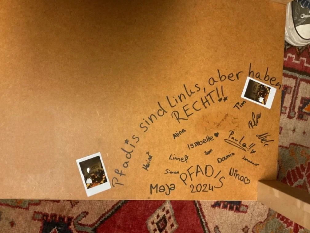 Un braunes Pappkarton mit handgeschriebenen Nachrichten und Unterschriften von mehreren Personen, darunter einige kleine Polaroid-Fotos, auf einem bunten Teppich.