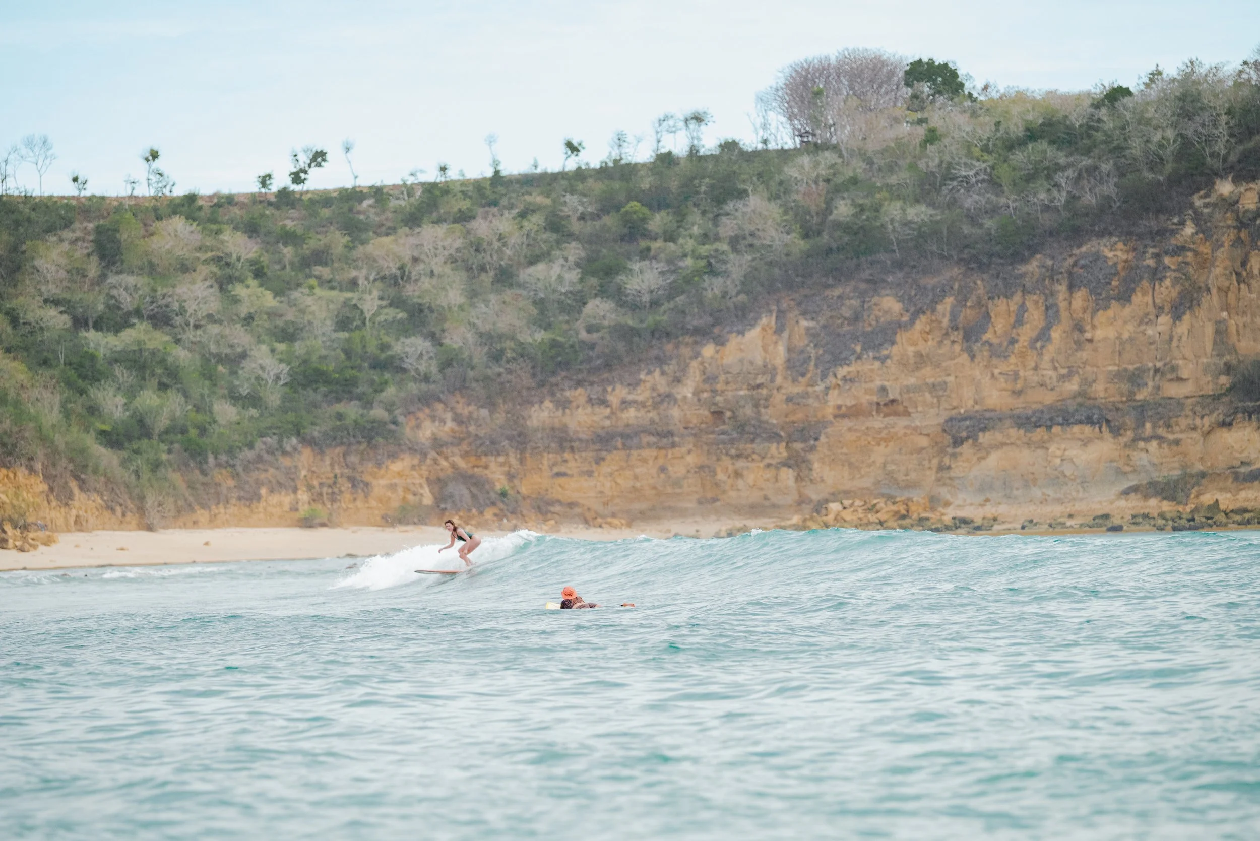 Surffaaja ajelee aaltoja rannikolla, taustalla pensasmainen mäki ja kalliot.