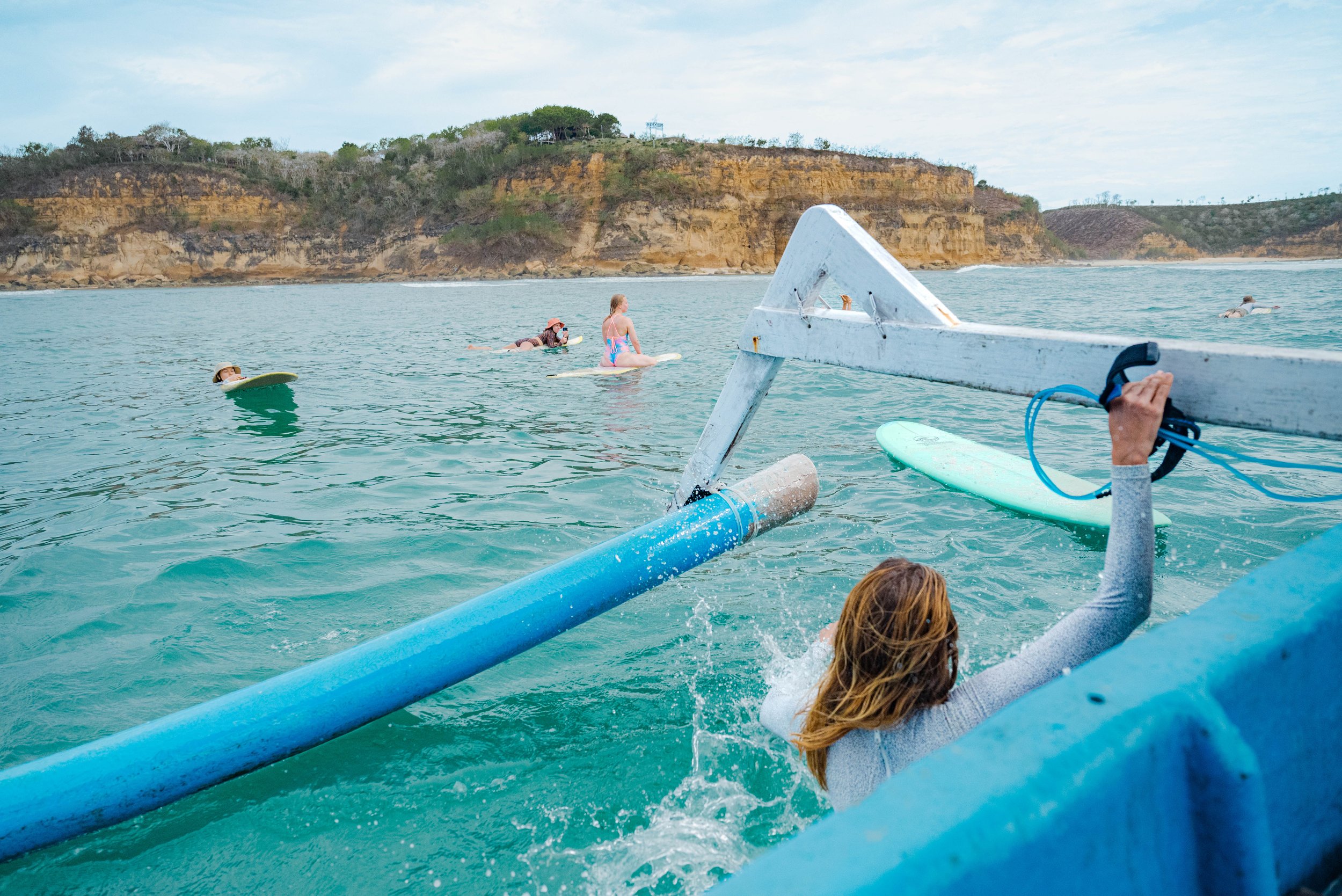 Uima-altaalla, nainen nousee pelastusliivistä sieppaamaan kajakkia tai leikkiväkeä, taustalla pulahtavia ihmisiä ja rannikko vuorineen.