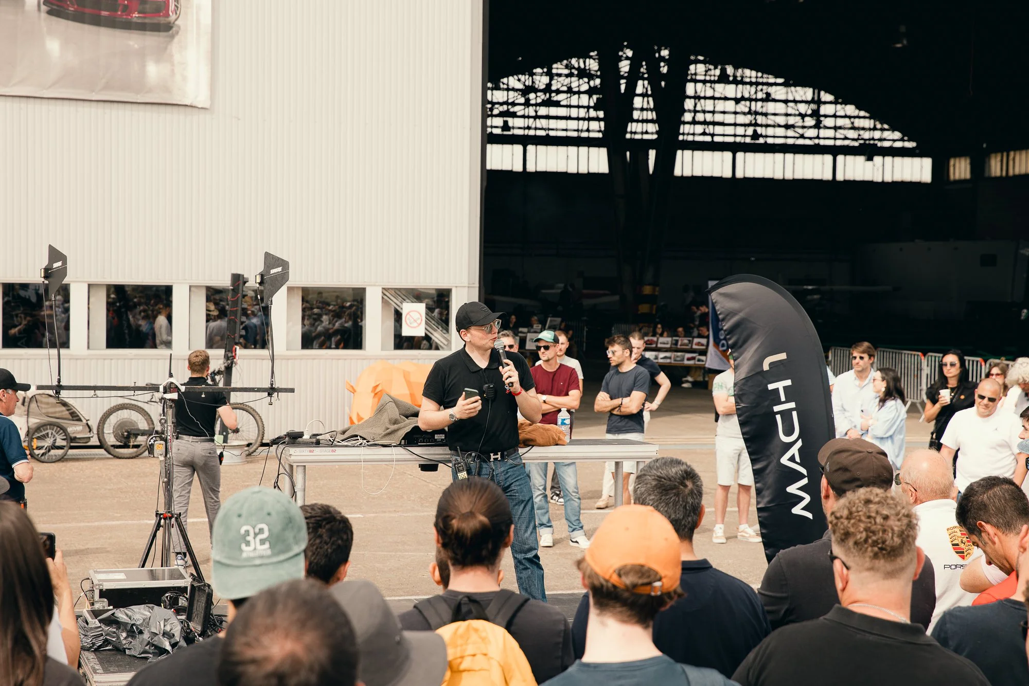 Un homme parle dans un micro lors d'un événement en plein air, entouré d'un public diversifié, à l'intérieur d'une structure comme un hangar, avec des voitures de sport en arrière-plan.
