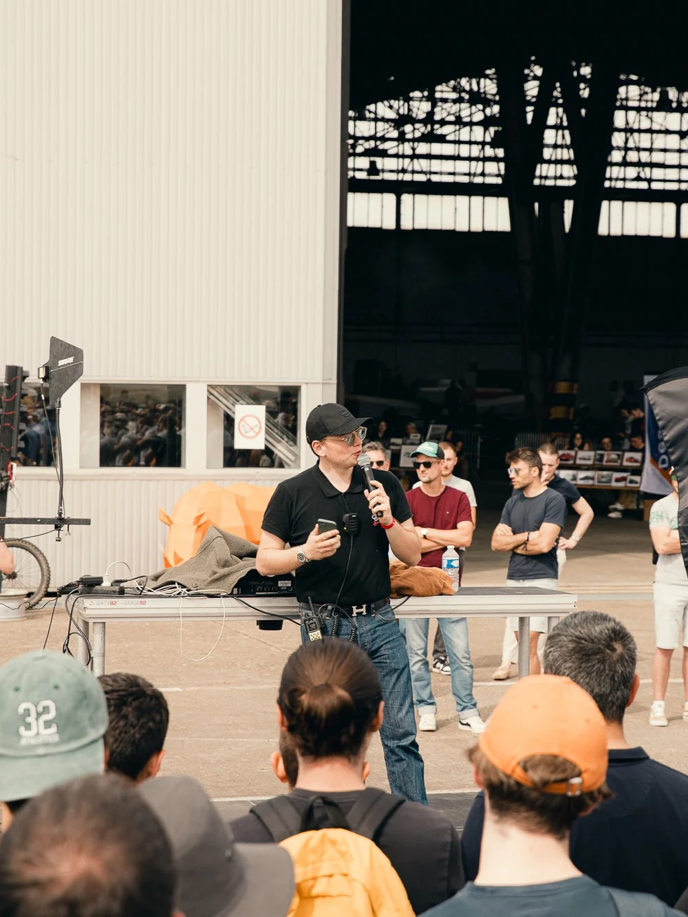 Le fondateur des Autophiles parle lors d'un événement public organisé à l'intérieur d'un bâtiment industriel, avec des centaines de personnes à l'écoute, certains portant des casquettes et des lunettes de soleil, service d'événementiel.