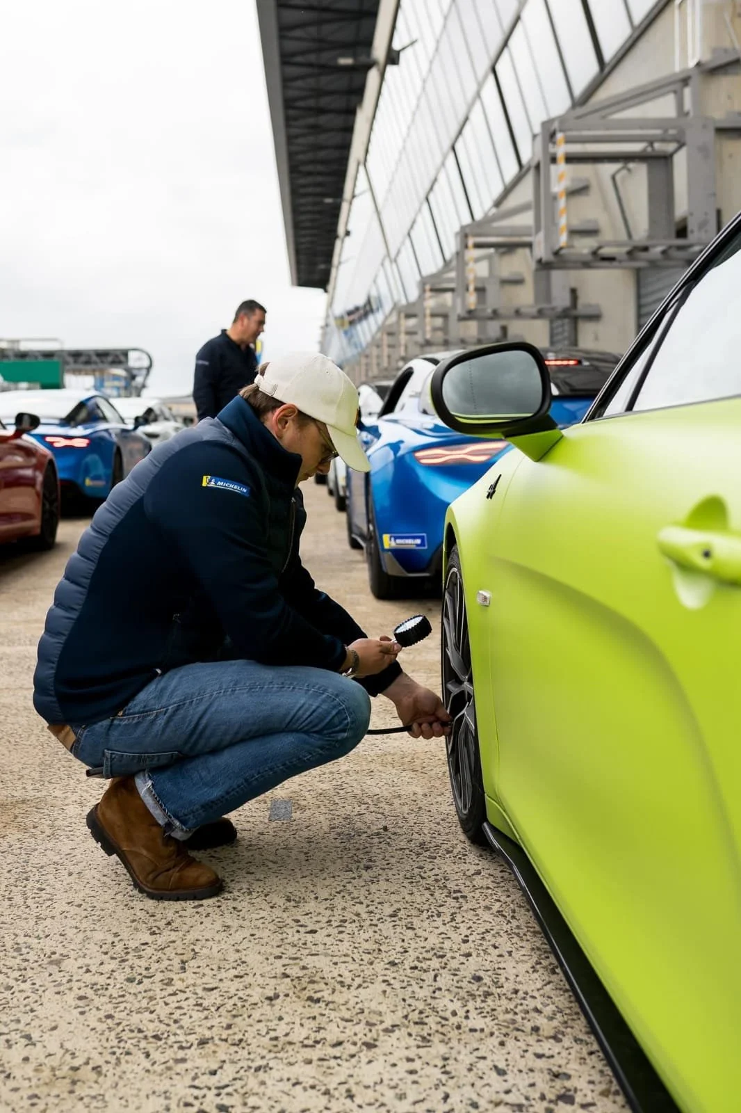 Le fondateur des Autophiles en jean, bottes de travail, veste foncée et casquette blanche vérifie la pression des pneus d'une voiture de course verte à l'aide d'un manomètre dans un garage de course pour illustrer le service d'expertise.