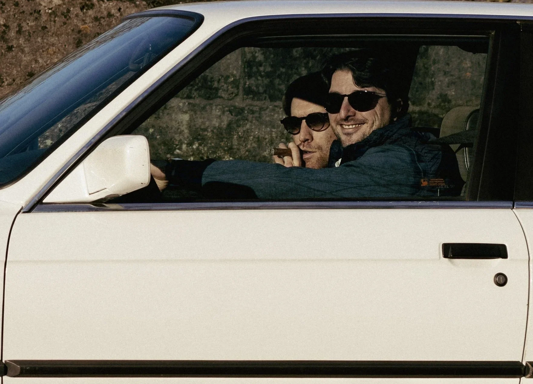 Deux hommes portant des lunettes de soleil dans une voiture blanche, l'un au volant et l'autre assis à côté, tous deux souriants, dans un environnement extérieur lors d'un rallye automobiles organisé par le club des Autophiles.