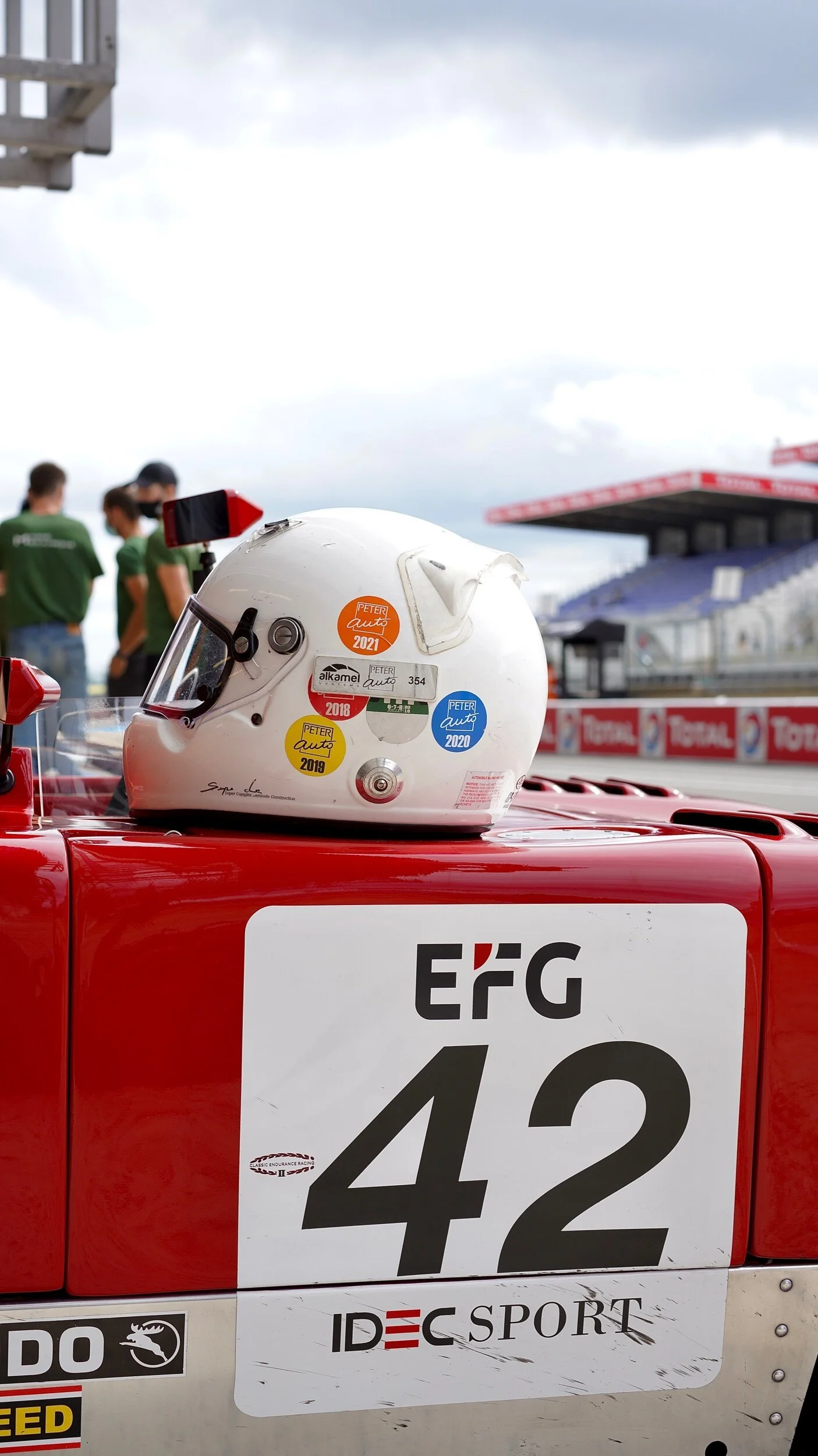Casque de course blanc avec des stickers indiquant différentes années et une voiture de course rouge avec le numéro 42 et le logo ERC, dans un environnement de circuit automobile organisé par les Autophiles.