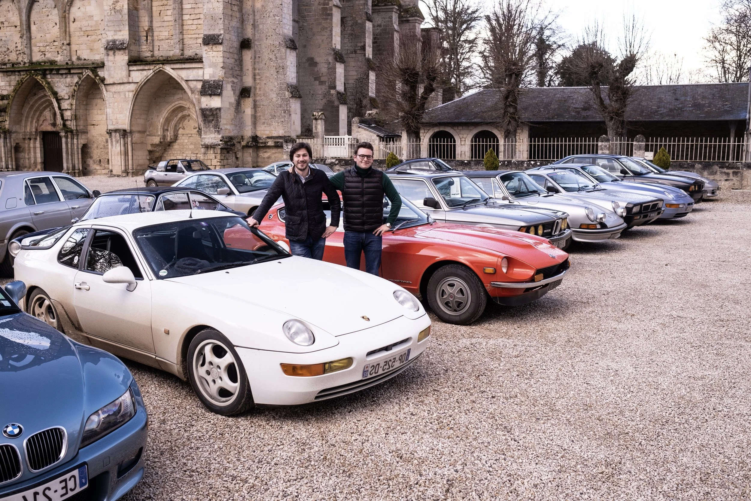Deux hommes posent devant un rassemblement de voitures de sport et collection, avec une église ancienne en arrière-plan pour représenter le club automobile Autophiles.