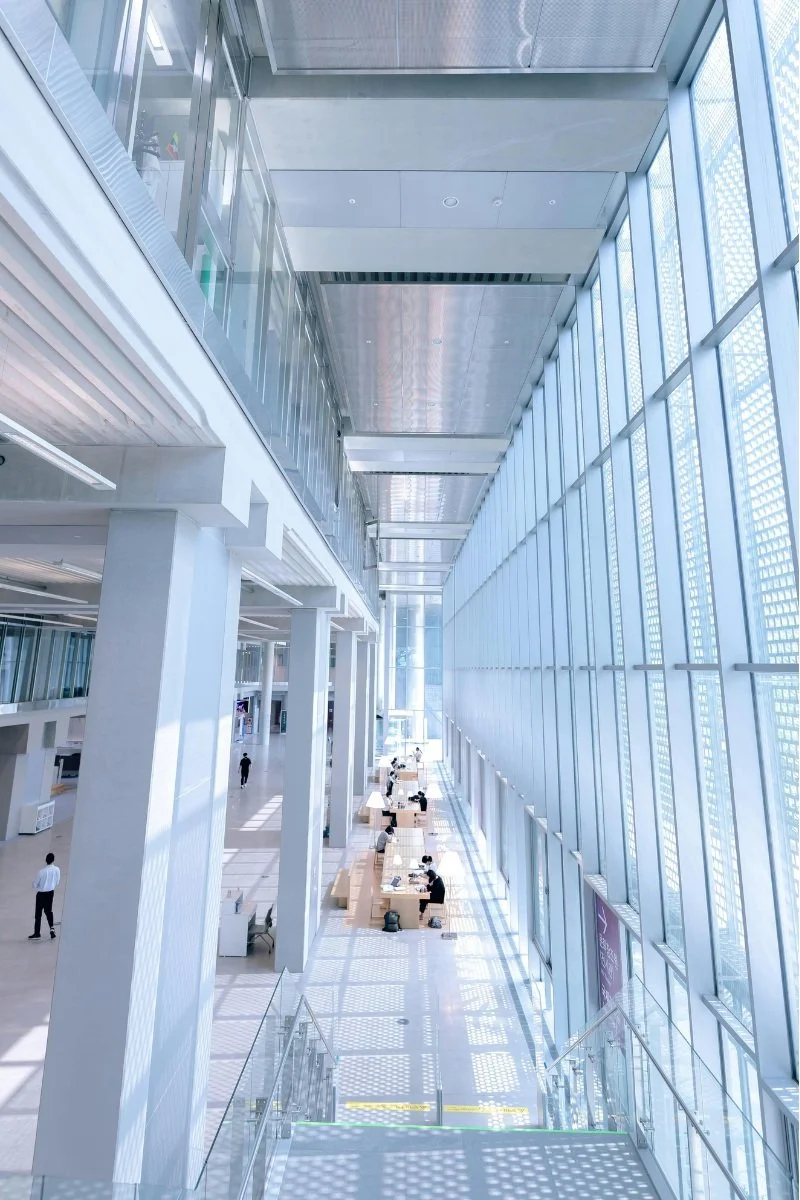 Intérieur lumineux d’un bâtiment moderne avec une verrière imposante, des colonnes blanches et un espace de travail partagé. Des personnes sont assises à des bureaux, profitant de la lumière naturelle.