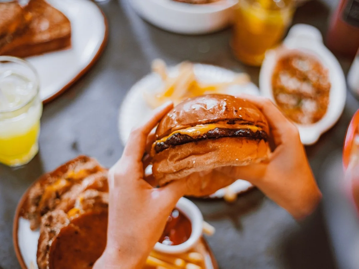 Personne qui tient un burger dans ses mains. La table est remplie de nourriture.