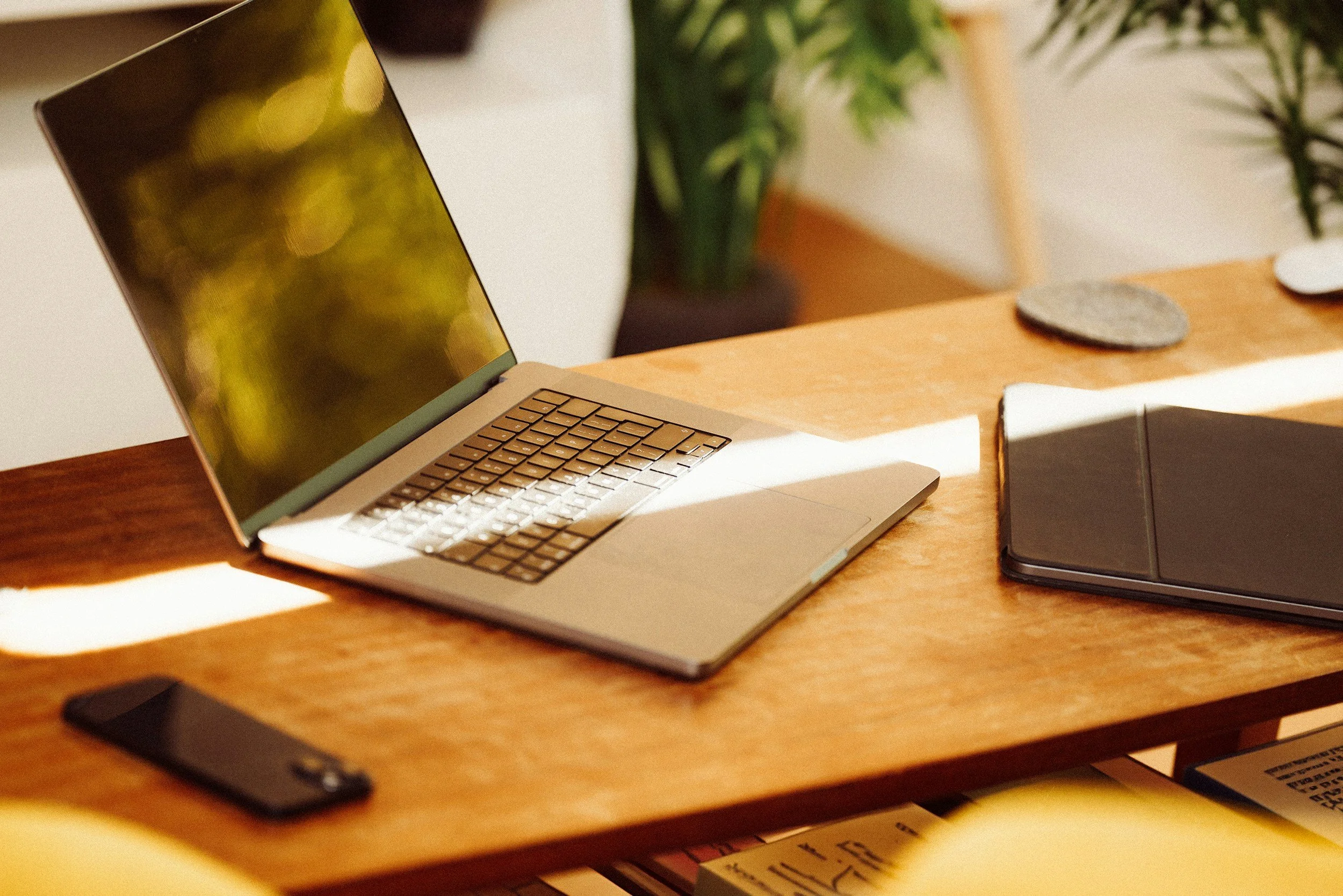 A wooden desk with a silver laptop, a smartphone, and a black closed laptop with stones and plants in the background.