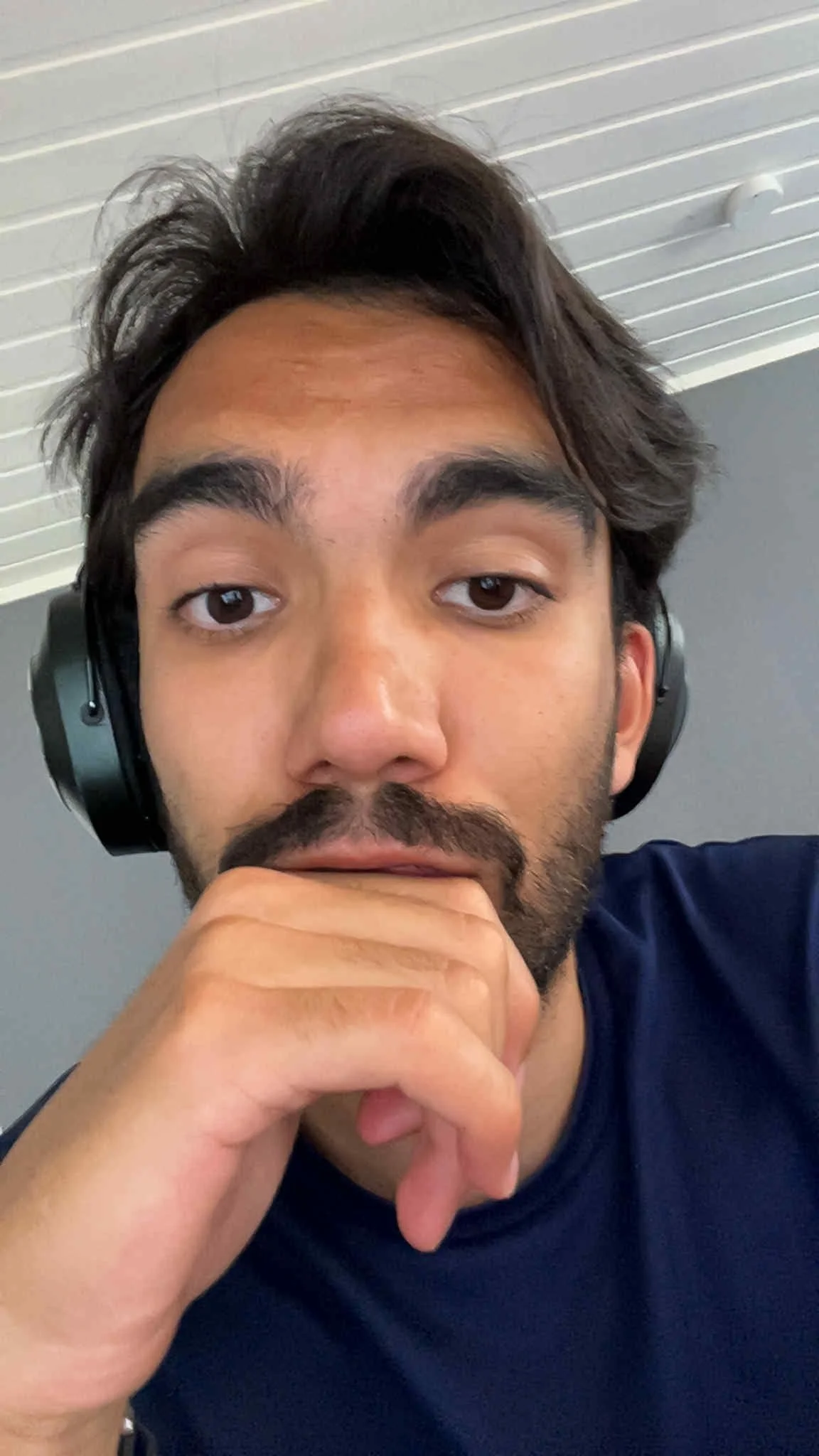 Close-up of a young man with dark hair, wearing headphones, resting his chin on his hand, looking directly at the camera.