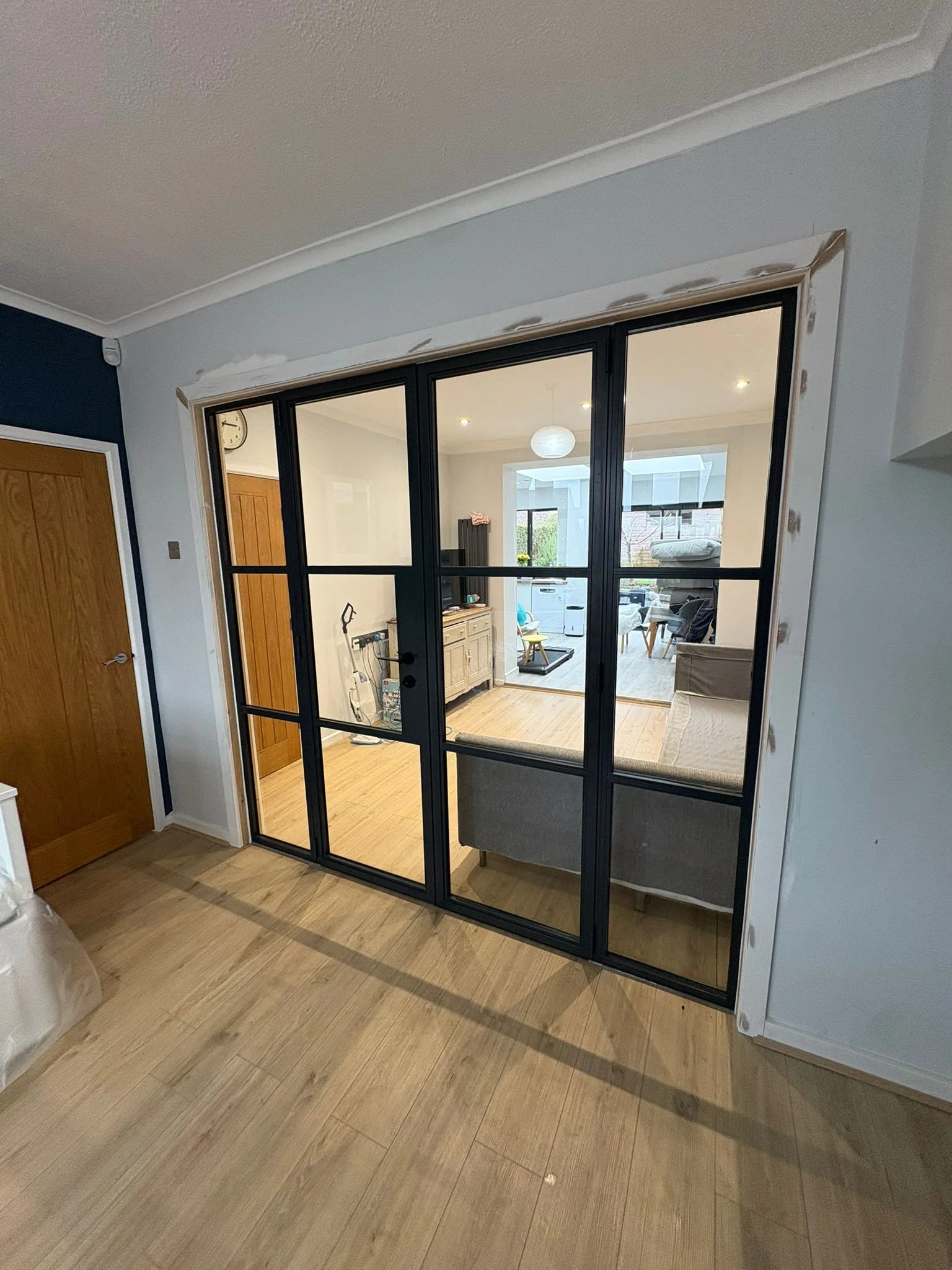View of a glass door with black metal frame leading to a living room with wooden floors, a gray sofa, and a white cabinet, with additional furniture and decor visible inside.