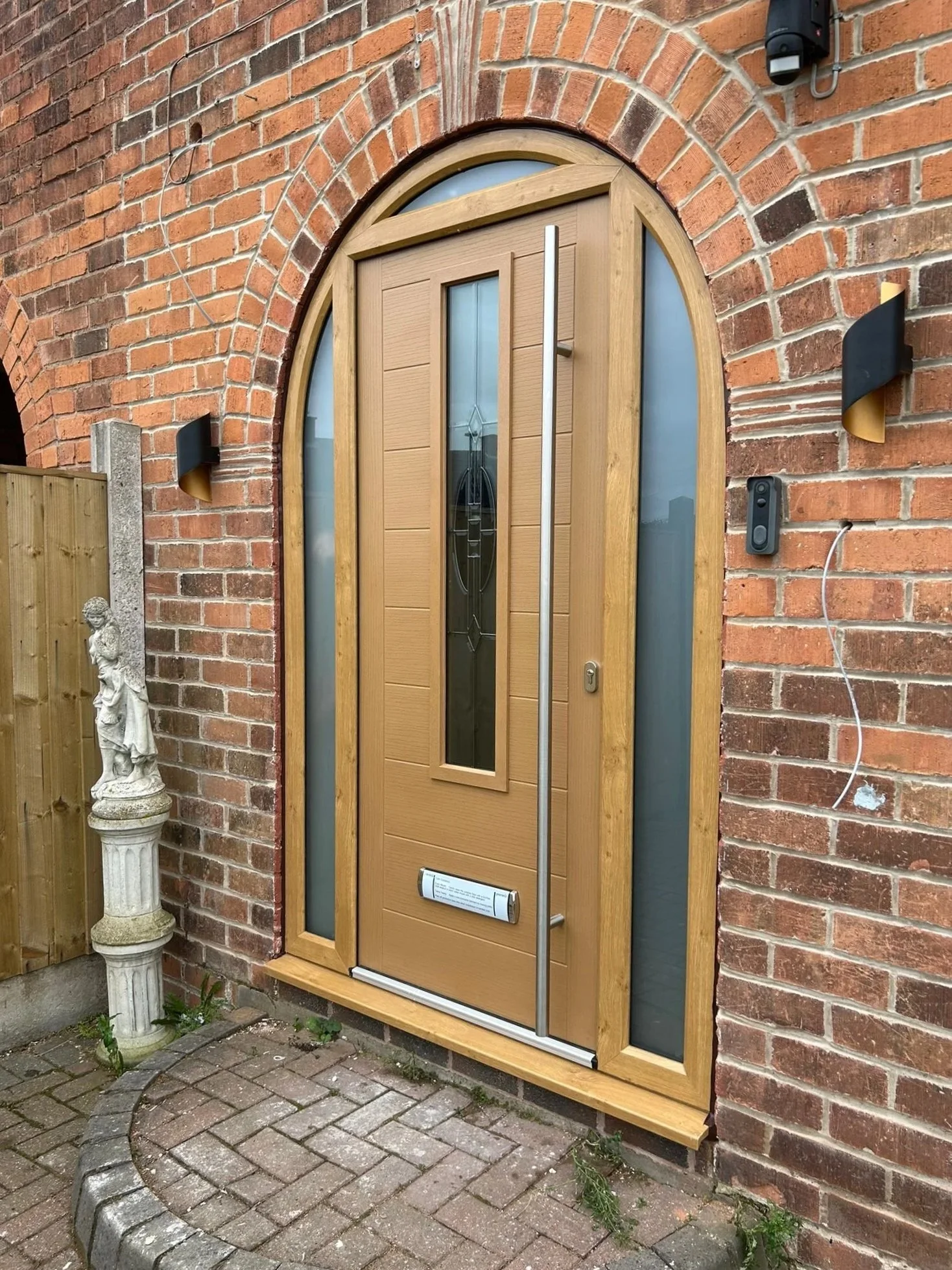 A wooden front door with a vertical glass insert, set in a brick wall. The door has a modern, sleek handle, and is surrounded by two narrow glass panels. There are two contemporary wall sconces on either side of the door, a camera or intercom to the right, and a statue on a pedestal near the entrance. The ground is paved with bricks and concrete.