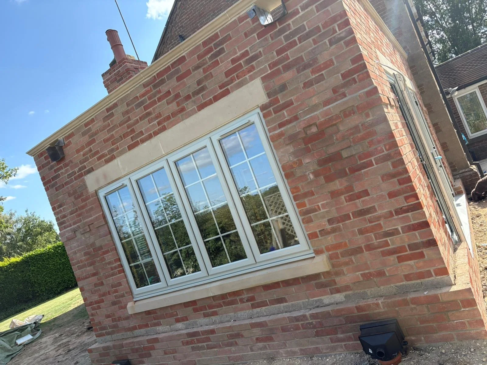 A brick house with large white-framed windows, a chimney, and a neighboring house visible in the background, set against a bright blue sky.