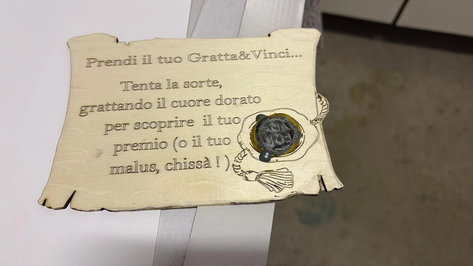 Cartello di legno con scritta in italiano che invita a prendere un biglietto e tentare la fortuna graffiando il cuore dorato. C'è anche un graffietto dorato con una capsula di moneta sopra.