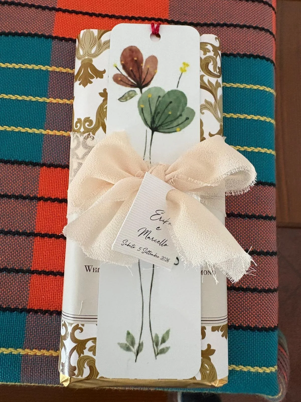 Confezione regalo decorata con un fiocco in tessuto crepe di color avorio. Sul pacchetto c'è un cartoncino con un disegno di due foglie di fiori in stile acquerello, una marrone e una verde chiaro, con piccoli fiori gialli. Su un'etichetta legata al 