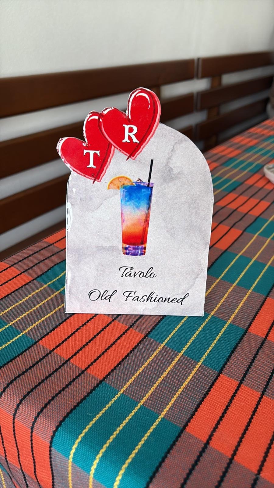 Segmento pubblicitario con immagine di un cocktail multicolore decorato con una fetta di limone e una cannuccia, con due cuori rossi sopra con le lettere T e R. Scritto 'Tavolo Old Fashioned'.