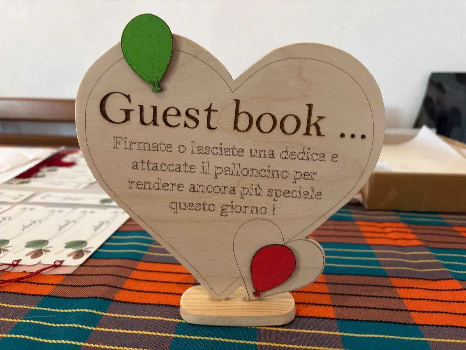 Segnaposto di legno a forma di cuore con scritta in italiano e palloncini decorativi verdi e rossi. Testo invita gli ospiti a firmare o lasciare un messaggio nel guest book per rendere speciale il giorno.