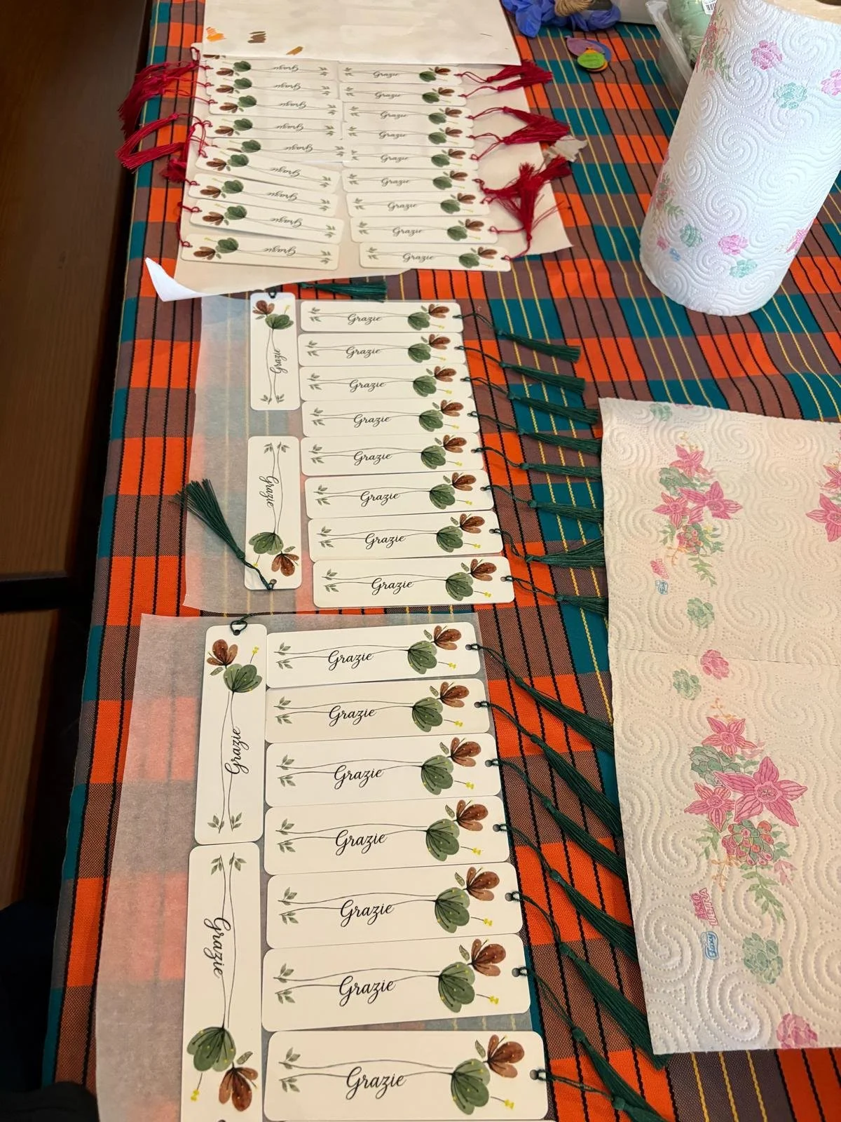 Tovaglioli di carta con decorazioni floreali rosa e verdi e scritte 'Grazie' posati su una tavola coperta con un tovagliato a quadri arancioni, verdi e blu. Sul tavolo ci sono anche alcuni cartellini con la stessa decorazione e la parola 'Grazie' scr