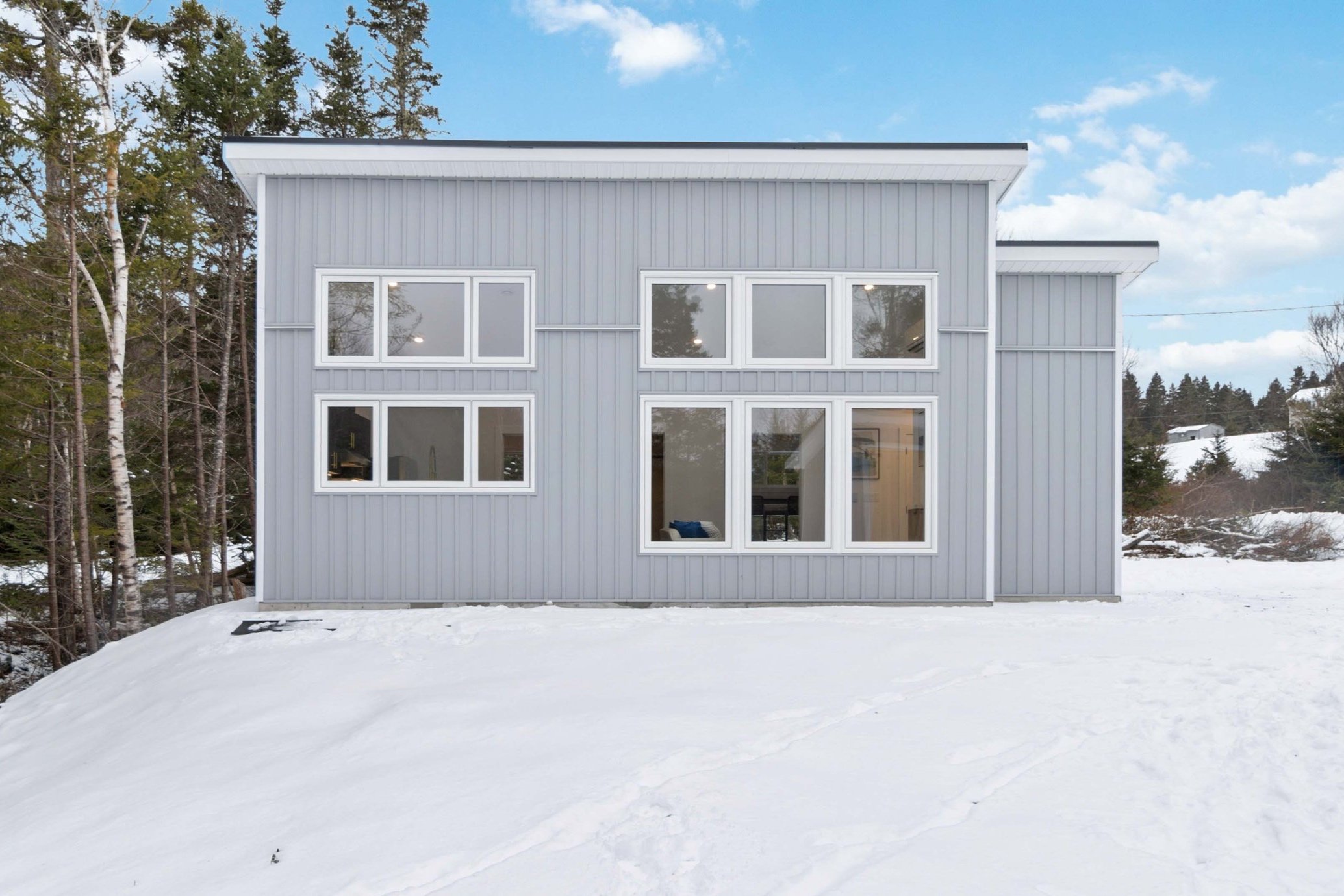 A modern building with gray siding and large white-framed windows, set against a snowy landscape with trees and a partly cloudy sky.