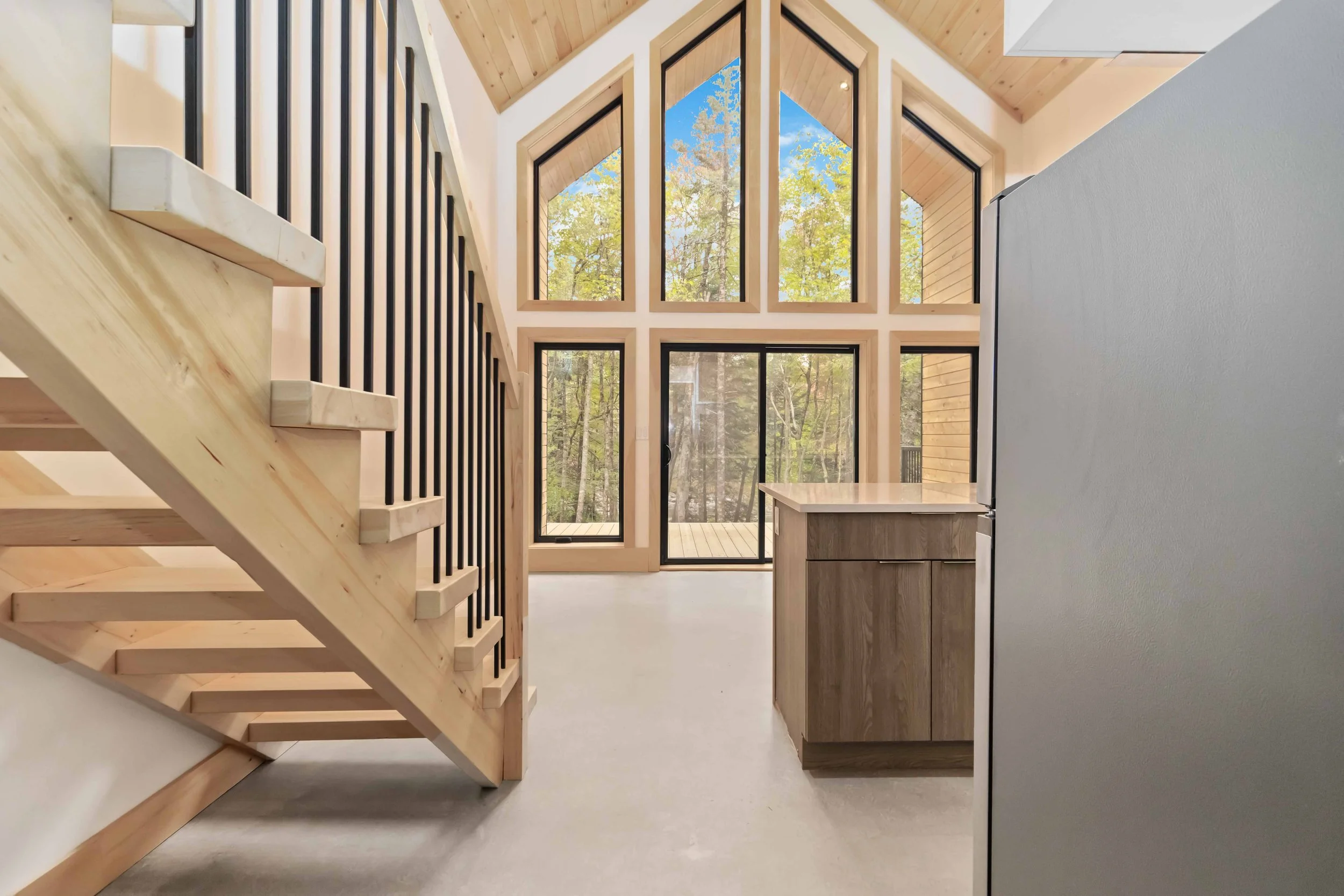 Interior of a modern house with a staircase on the left, large windows showing trees outside, and a kitchen area on the right with wooden cabinets.