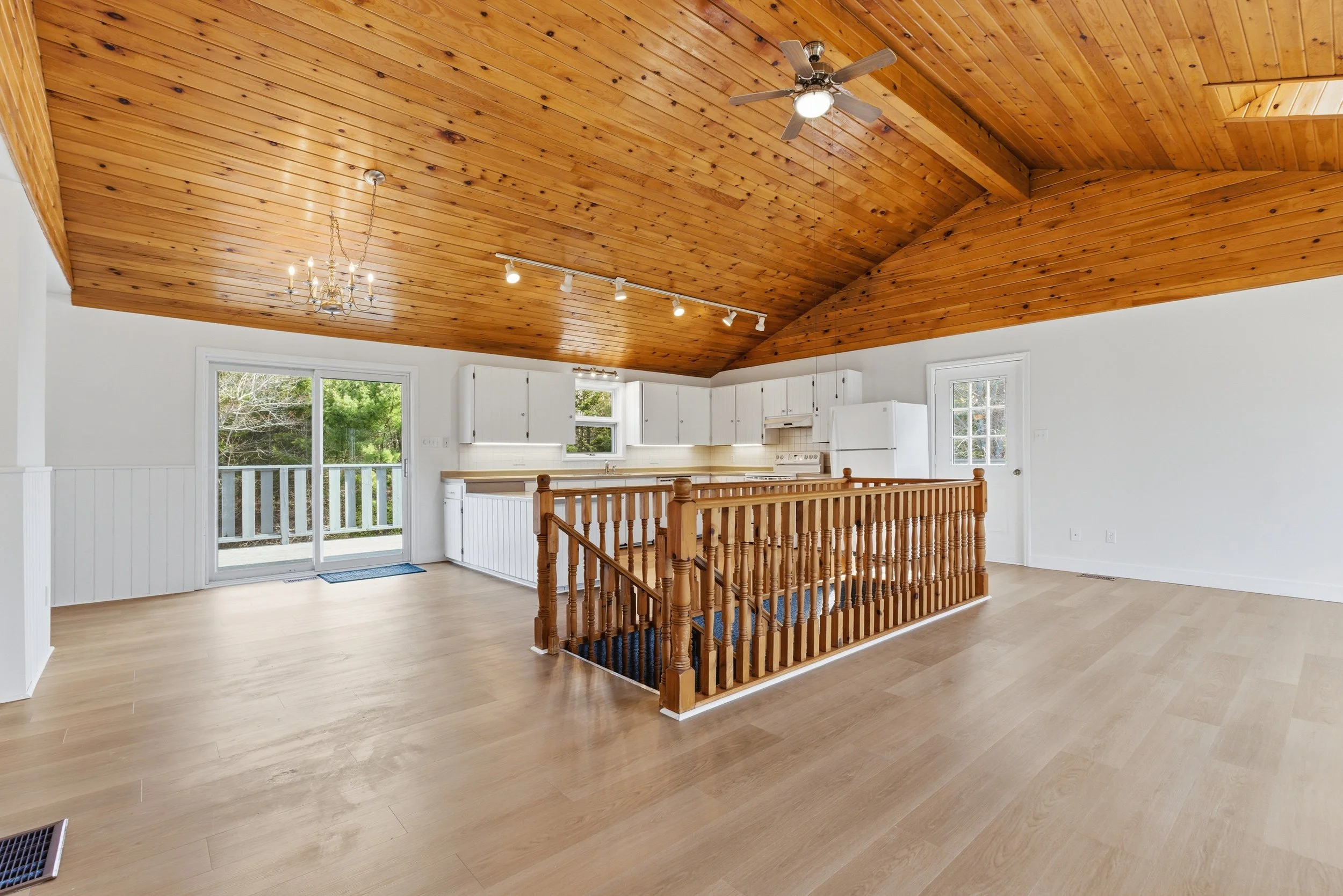 Living room area with a wood-paneled vaulted ceiling, ceiling fan, chandelier, large sliding glass door, white kitchen cabinets, and hardwood flooring.