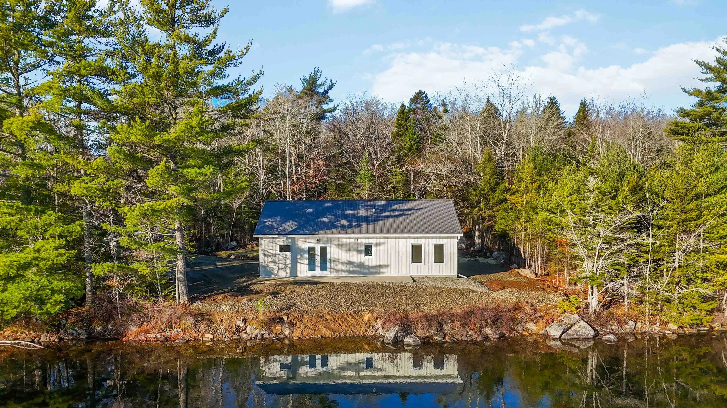 Small white house with a dark gray metal roof nestled among trees near a body of water, under a blue sky with some clouds.