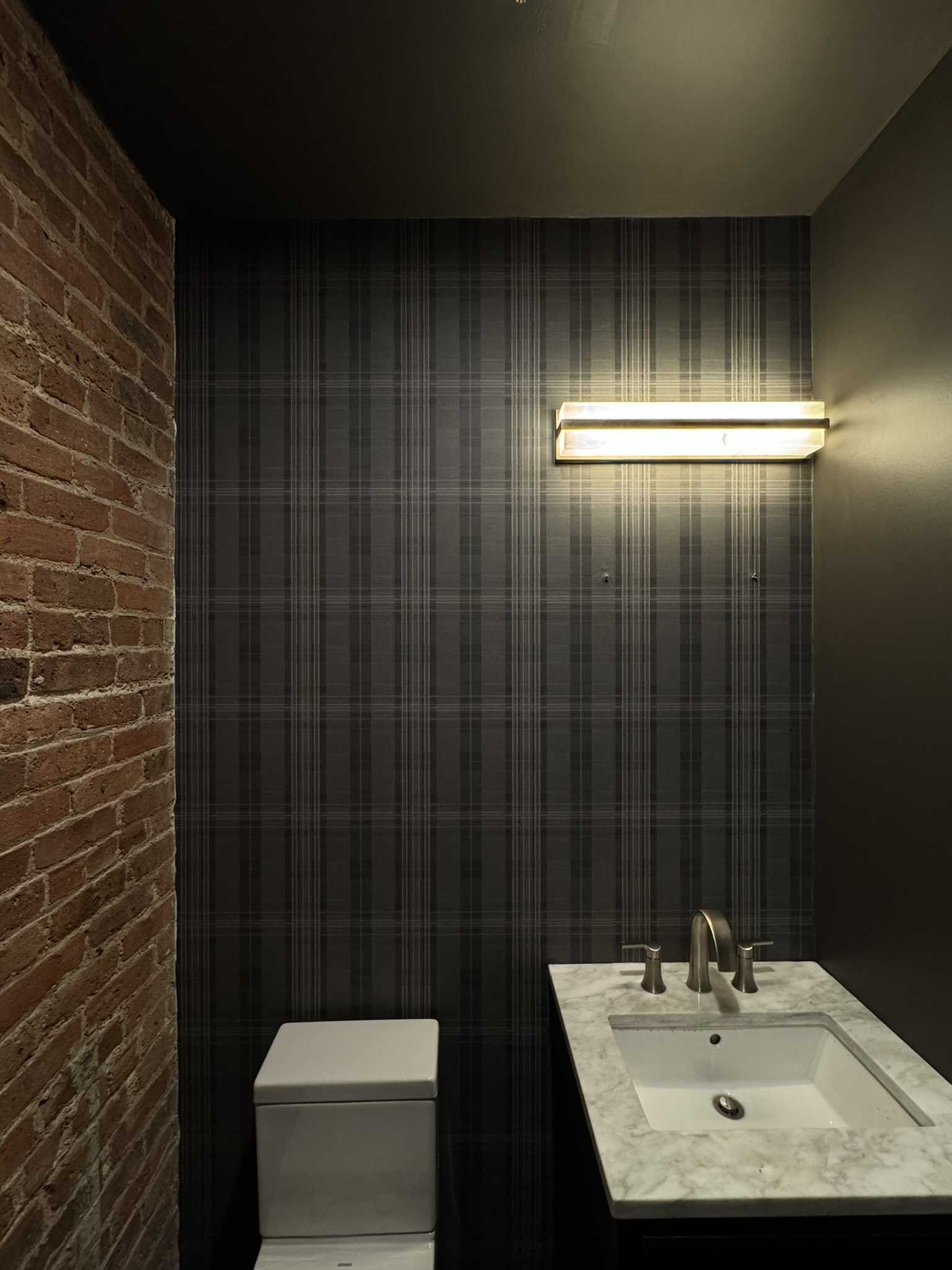 A bathroom with a brick wall on the left, brown plaid wallpaper on the back wall, and a dark green wall on the right. A wall-mounted light fixture is above the mirror. A white toilet with a closed lid is next to a marble countertop with a rectangular sink and a silver faucet.
