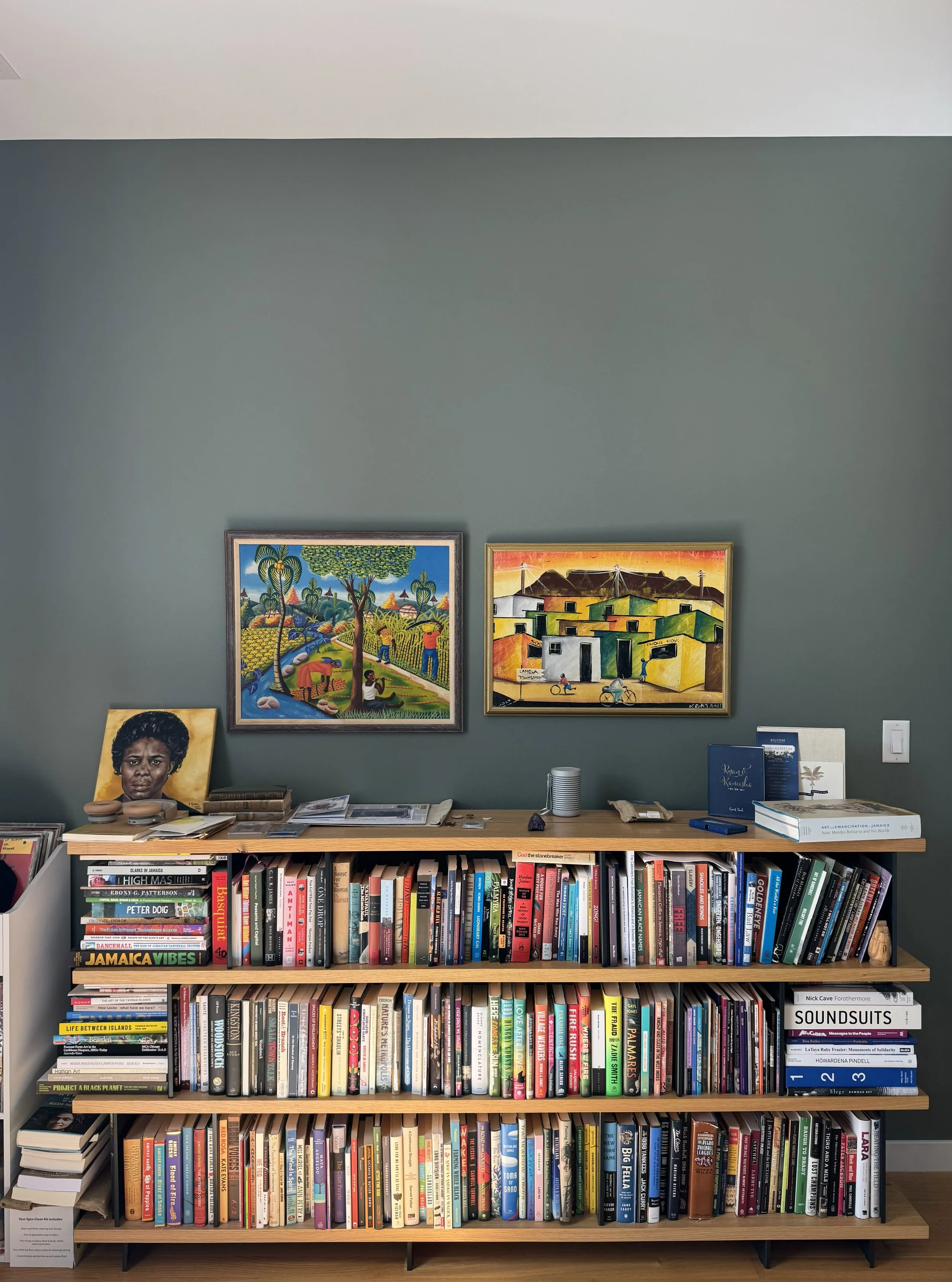 A wooden bookshelf filled with a wide variety of colorful books, with two art pieces hanging above it on a green wall. There are some objects like paper, a cup, and small items on top of the bookshelf.