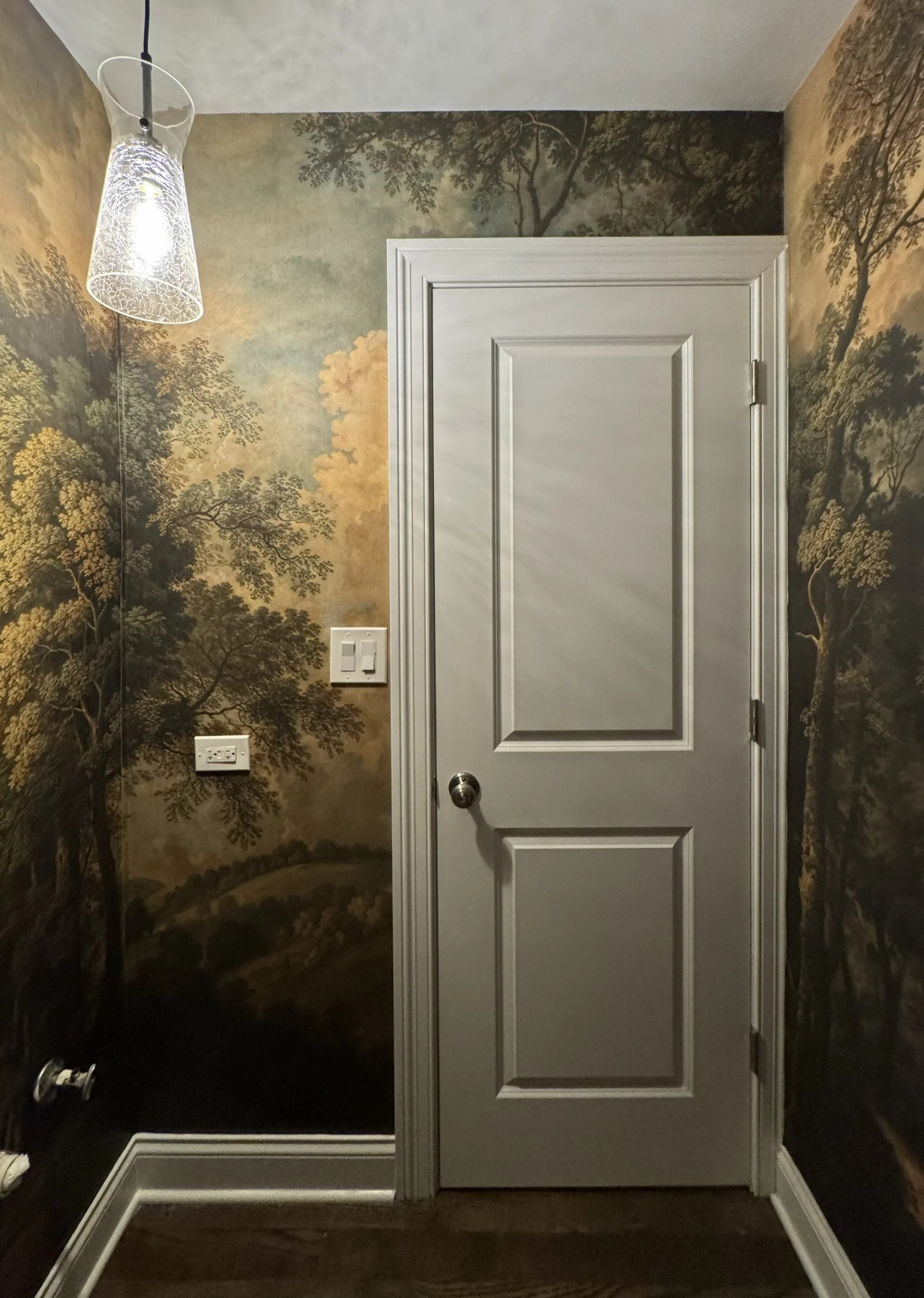 A room with wallpaper featuring trees and a landscape, a white door, a wall switch, and a floor vent.