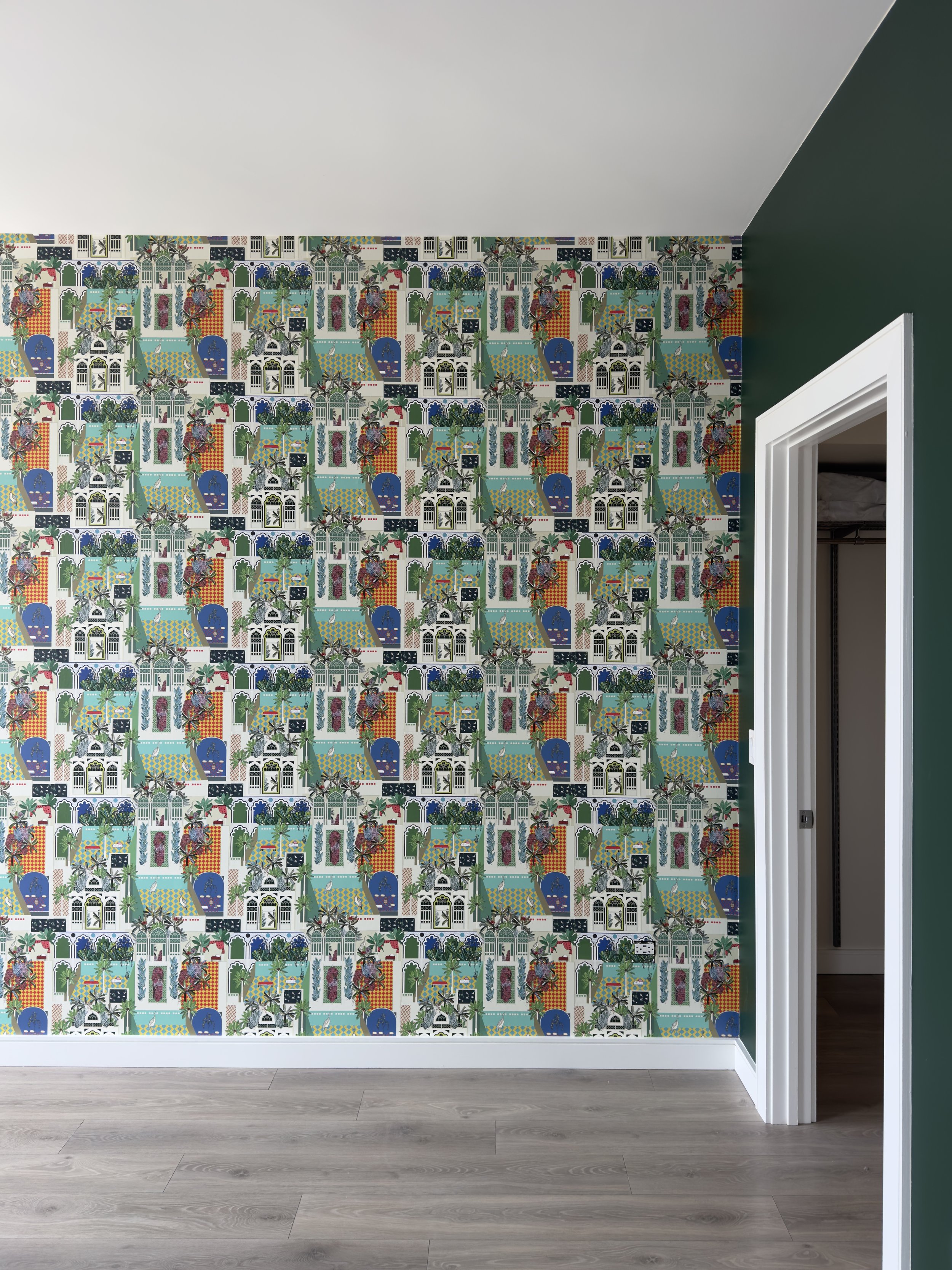 Interior wall with colorful patterned wallpaper featuring various geometric and botanical designs, next to an open doorway with white trim, and a light wood floor.
