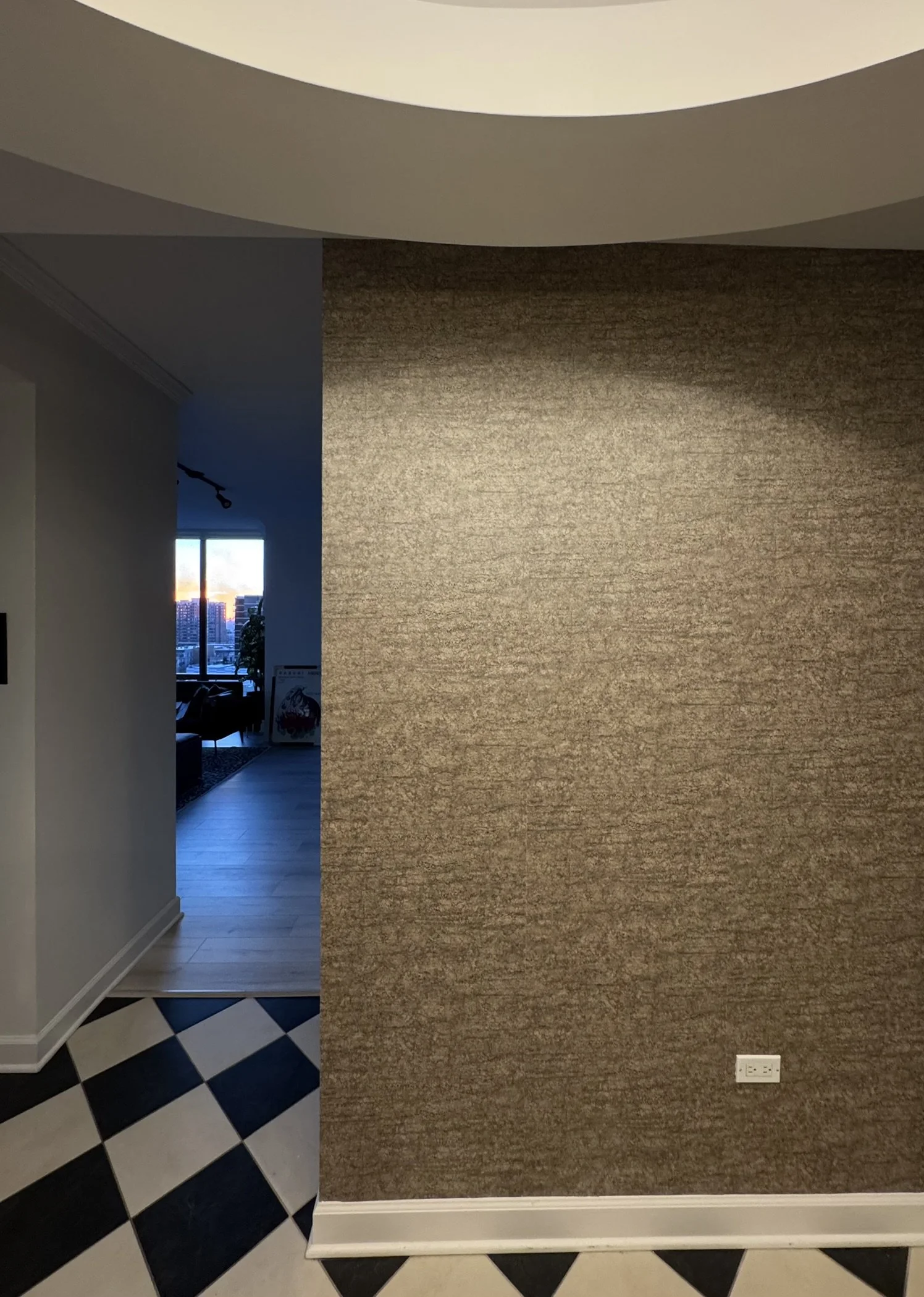 Interior view of a room showing a large textured brown wall with an electrical outlet at the bottom right. To the left, part of a patterned blue and white tile floor and a doorway opening to a brighter room with a window showing a cityscape at sunset.