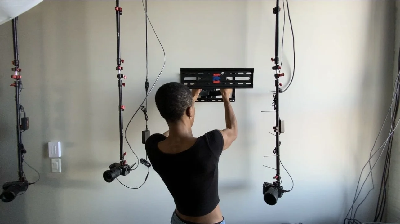 A woman hanging a black metal plate on a wall for a photo or video shoot, with lighting equipment on either side of her.