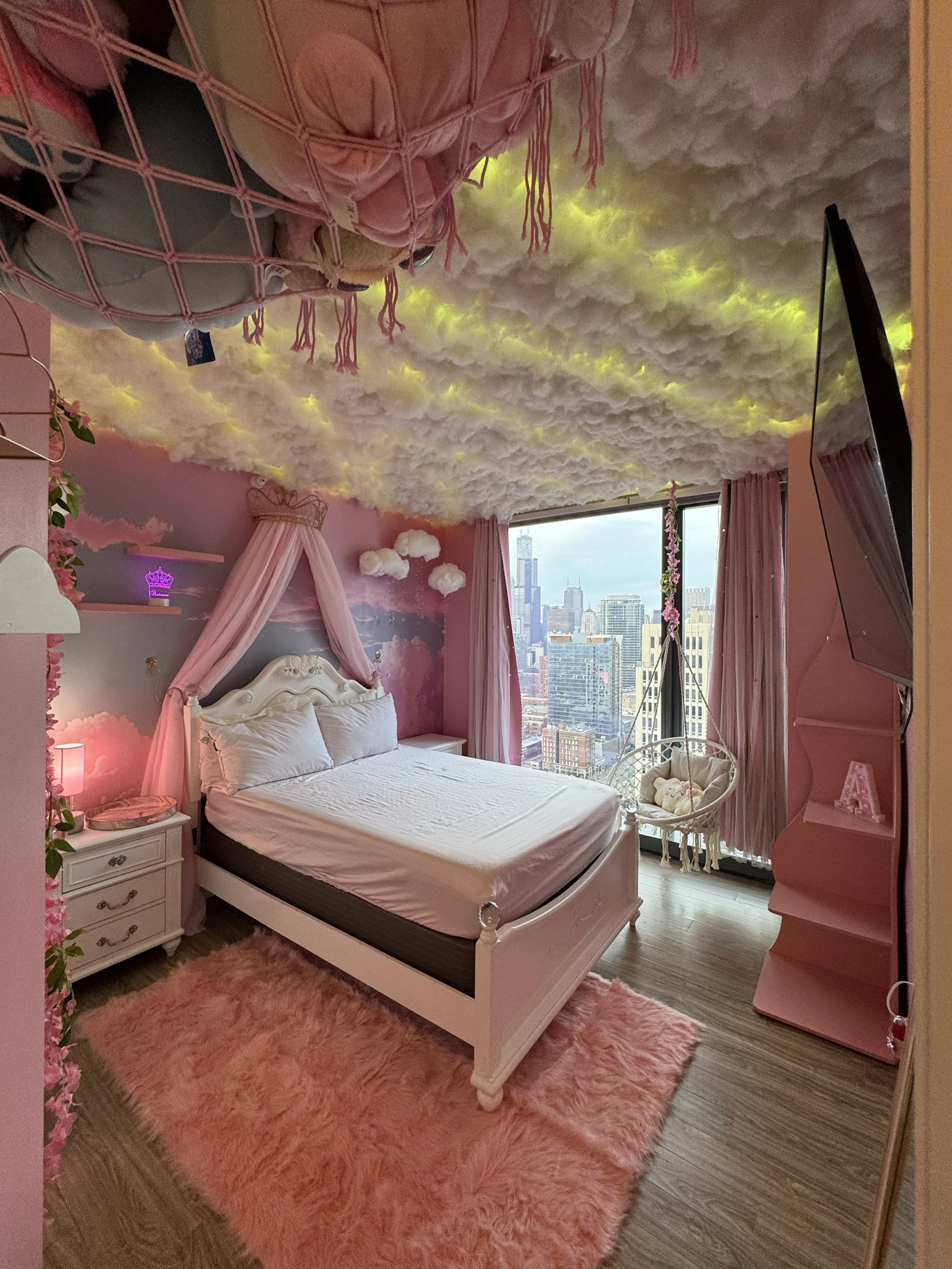 A bedroom with pink clouds on the ceiling and pink-themed decor. There is a white bed with a canopy and white bedding, a pink shaggy rug, a small white nightstand with drawers, and a window showing a city skyline. The room has pink curtains and a pink dresser, with decorative items like a teddy bear and a flower garland.