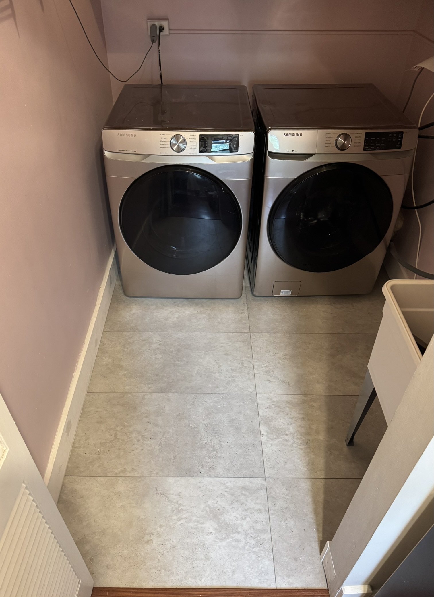 A laundry room with a washing machine and dryer side by side, a beige wall, and tile flooring.