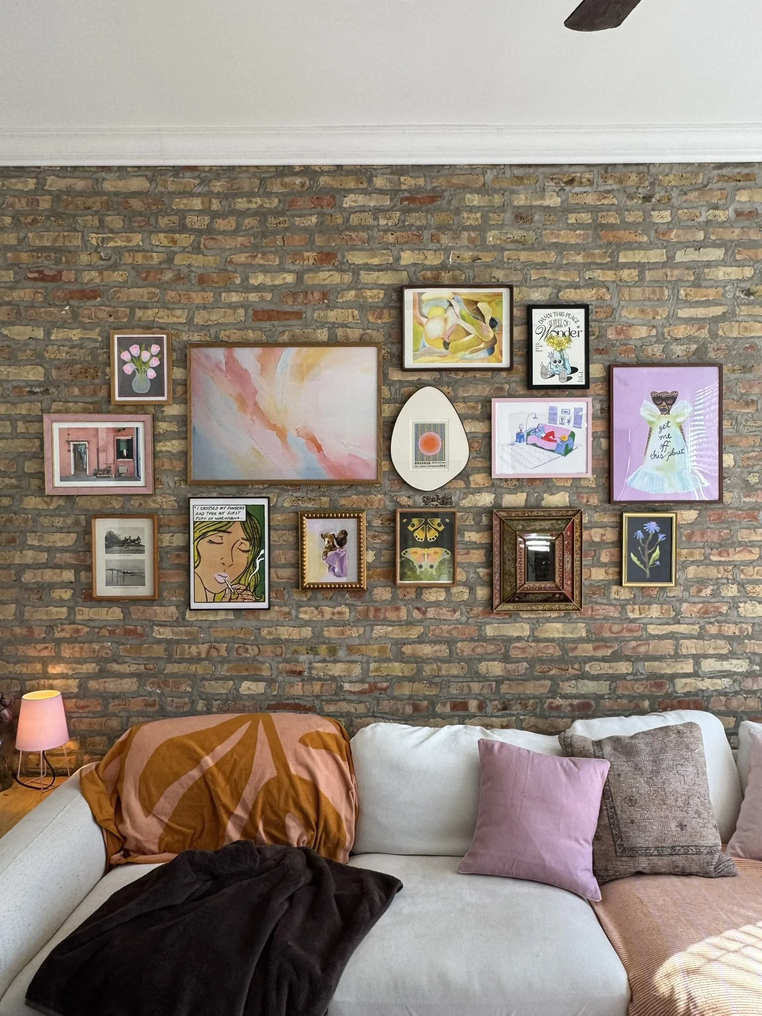 Living room with a white sofa, purple and brown pillows, a brown throw blanket, a pink pillow, a skirt, a bedside lamp, and a gallery wall with framed artwork and photographs on a brick accent wall.