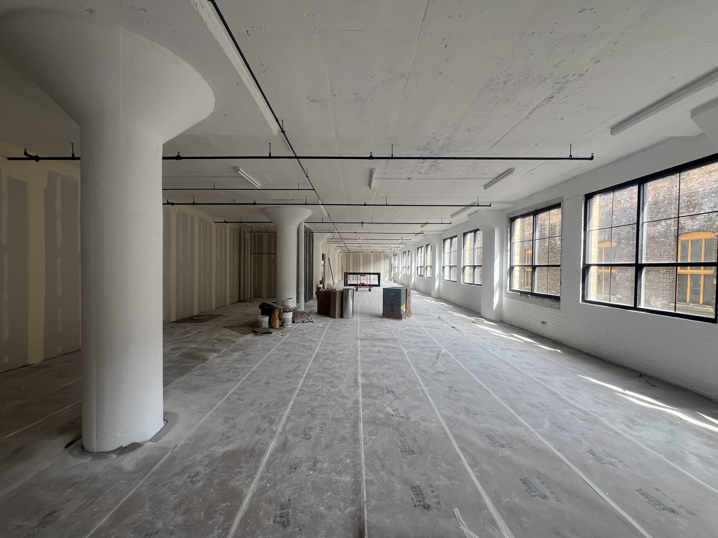 An unfinished interior space with large windows, visible construction materials, and exposed ceilings with pipes and lighting fixtures, indicating ongoing building or renovation work.
