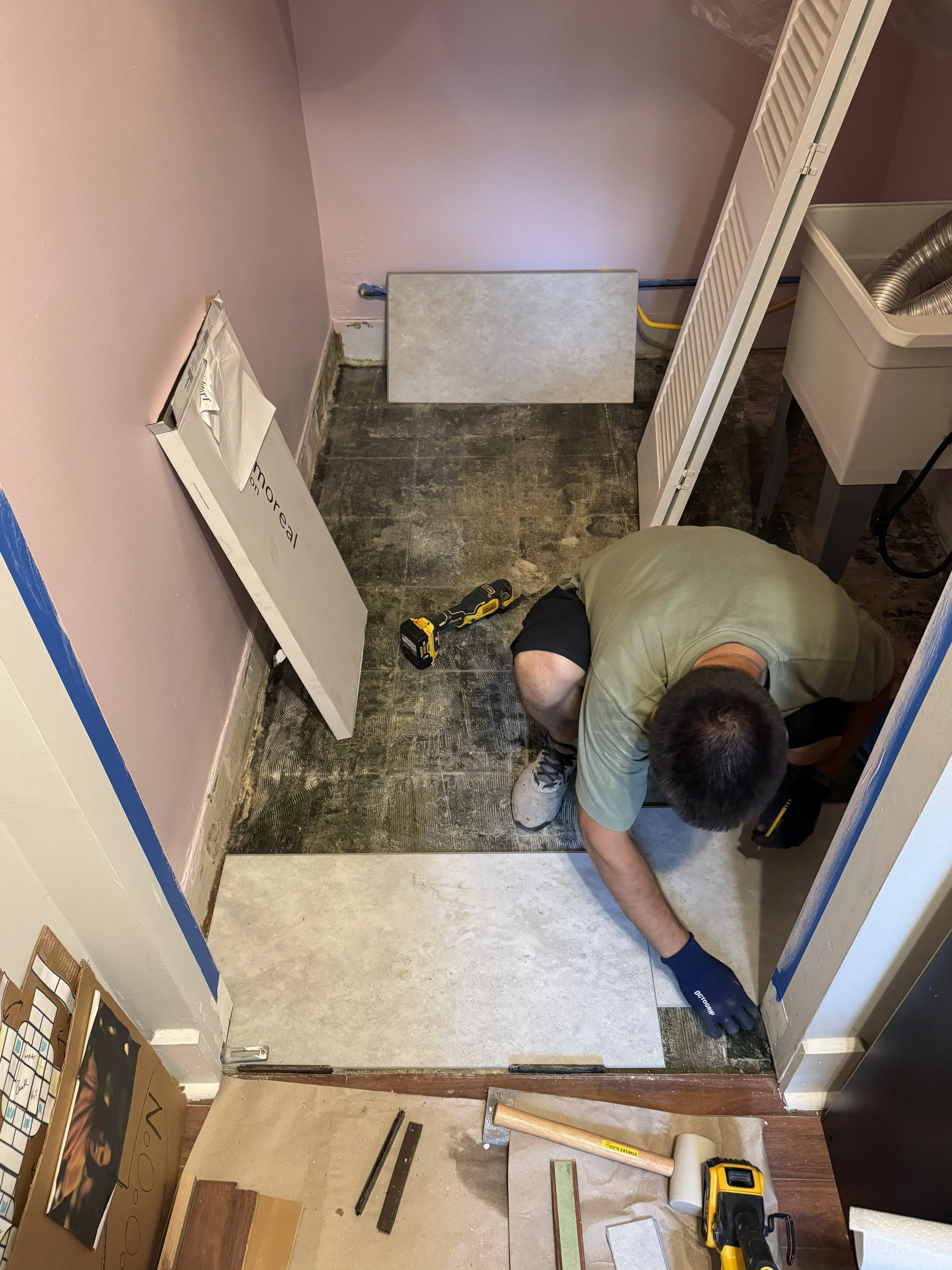 A person installing or working on flooring in a small room, with various tools and supplies around, including a drill, measuring tape, and tiles, while kneeling on the floor.