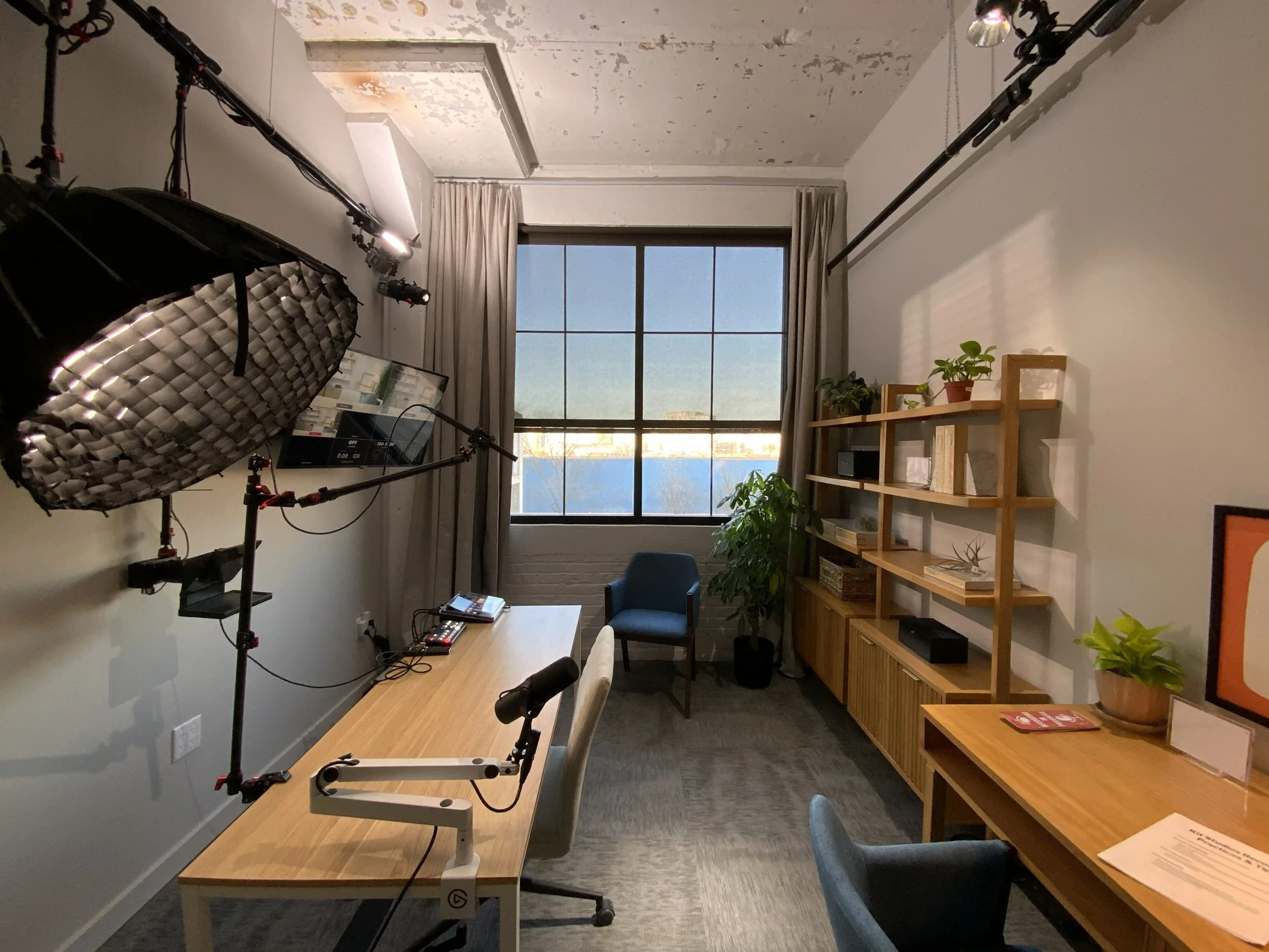 Indoor room with a large window, curtains, a wooden bookshelf with potted plants and books, a table with chairs, and professional lighting equipment.