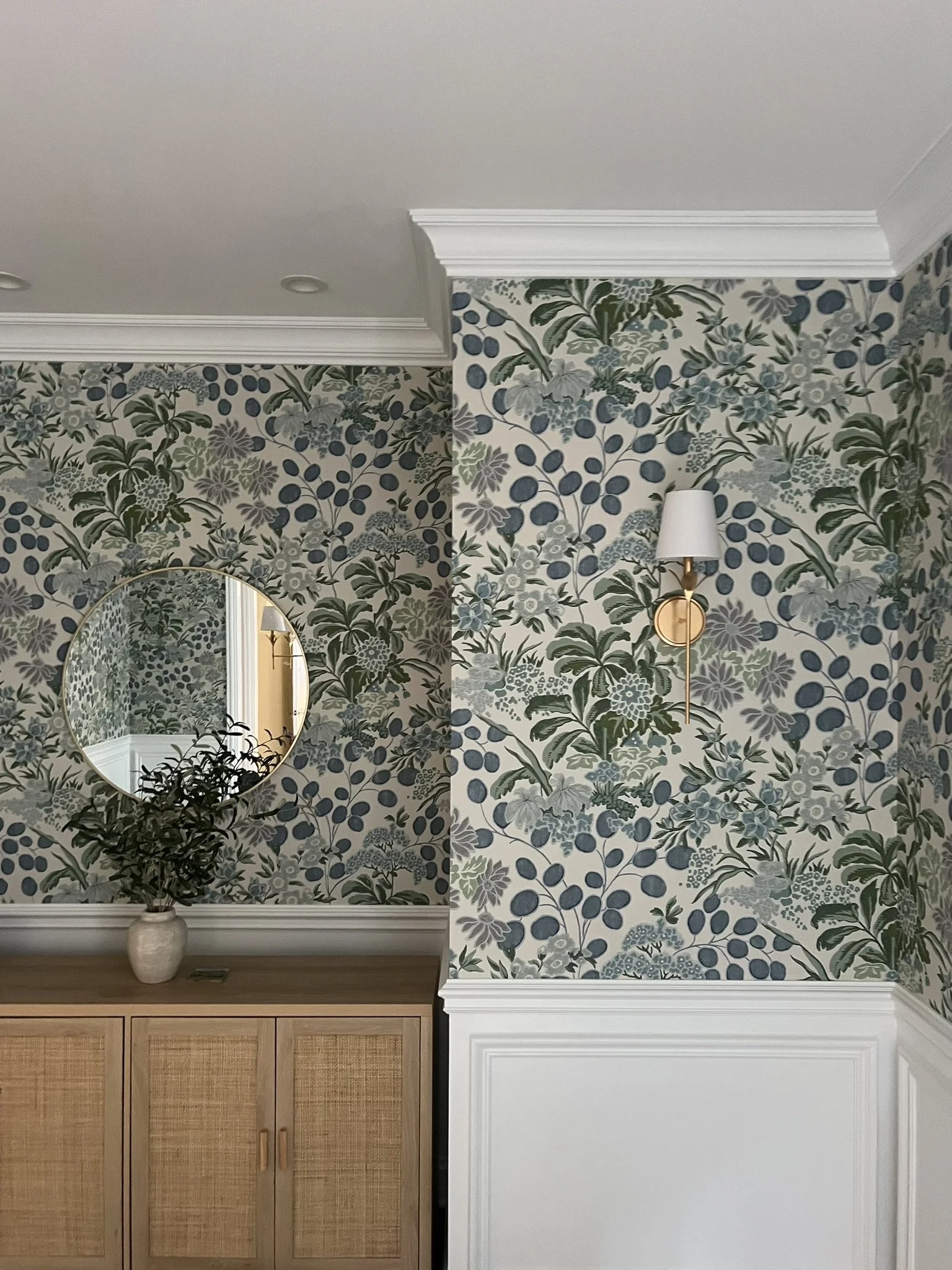 Interior view of a room with floral wallpaper, a wall sconce light, a round mirror, a vase with a plant, and wooden cabinets with rattan fronts.