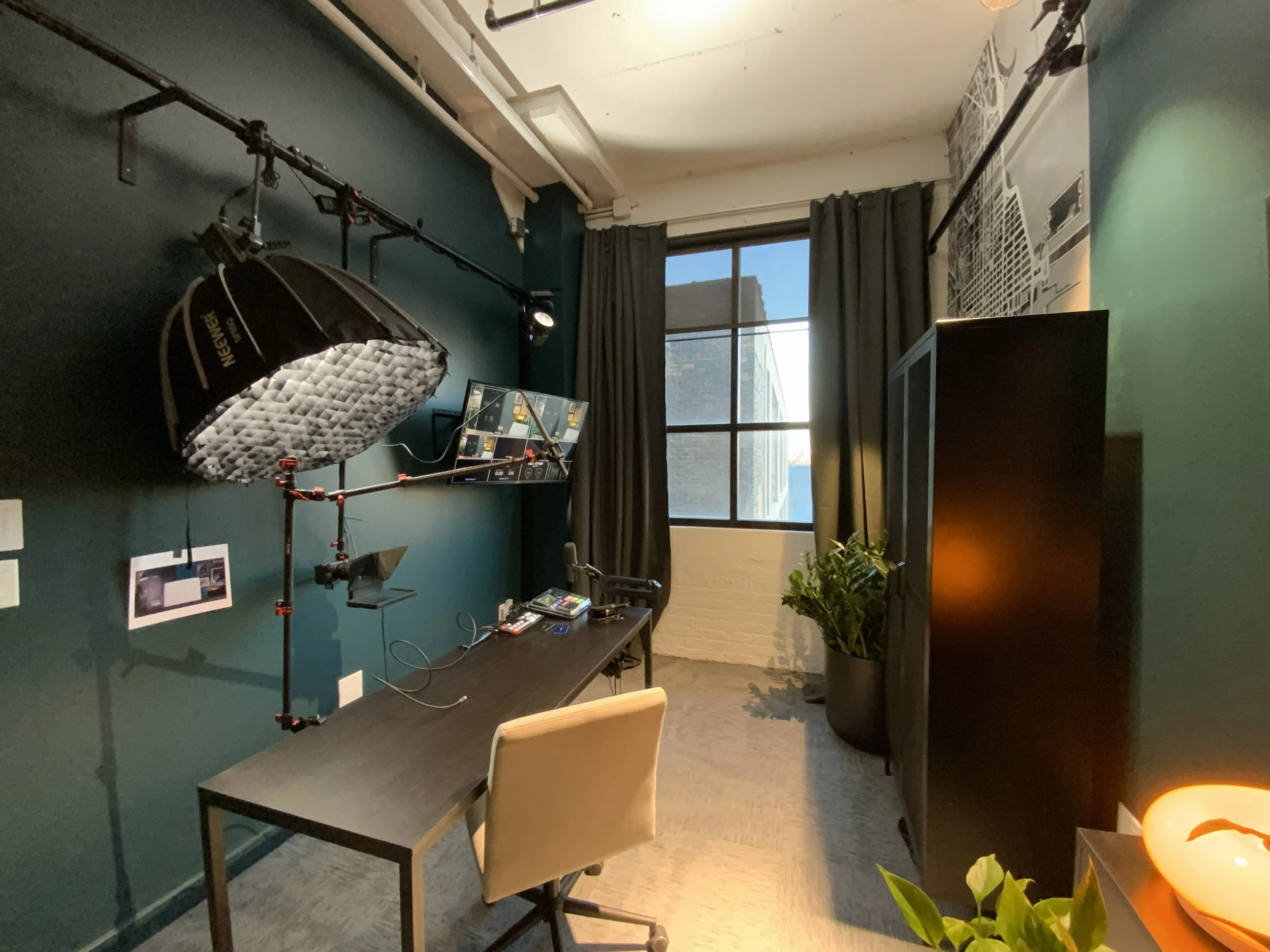 A room with a dark green wall decorated with photos, an overhead adjustable light, a window with dark curtains, and photography equipment including a softbox and camera settings displayed on a monitor.