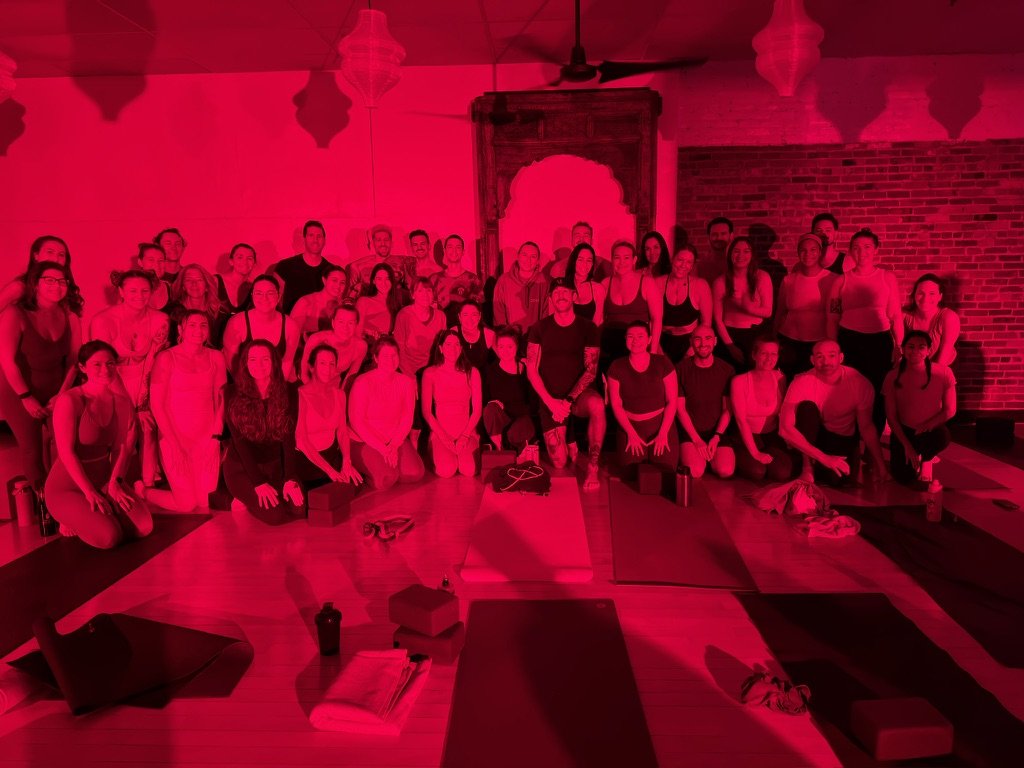 Group of people participating in a yoga class in a studio with brick wall and ornate door, illuminated with red lighting.
