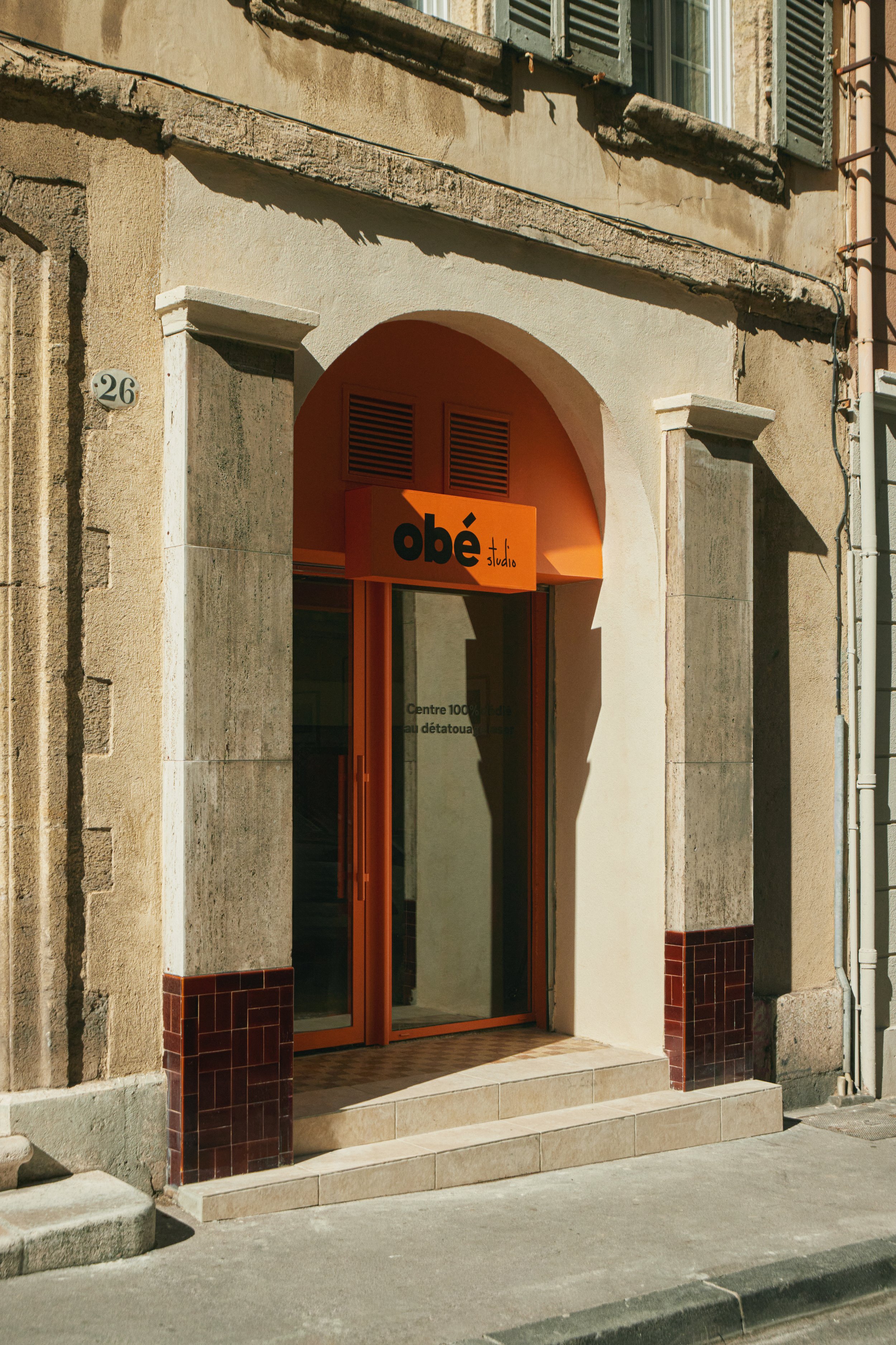 Façade d’un centre de détatouage laser à Marseille conçu par Local Club, agence d’architecture et design spécialisée dans les lieux de santé et commerces. Entrée marquée par une arche existante et une intervention contemporaine orange, créant une ide